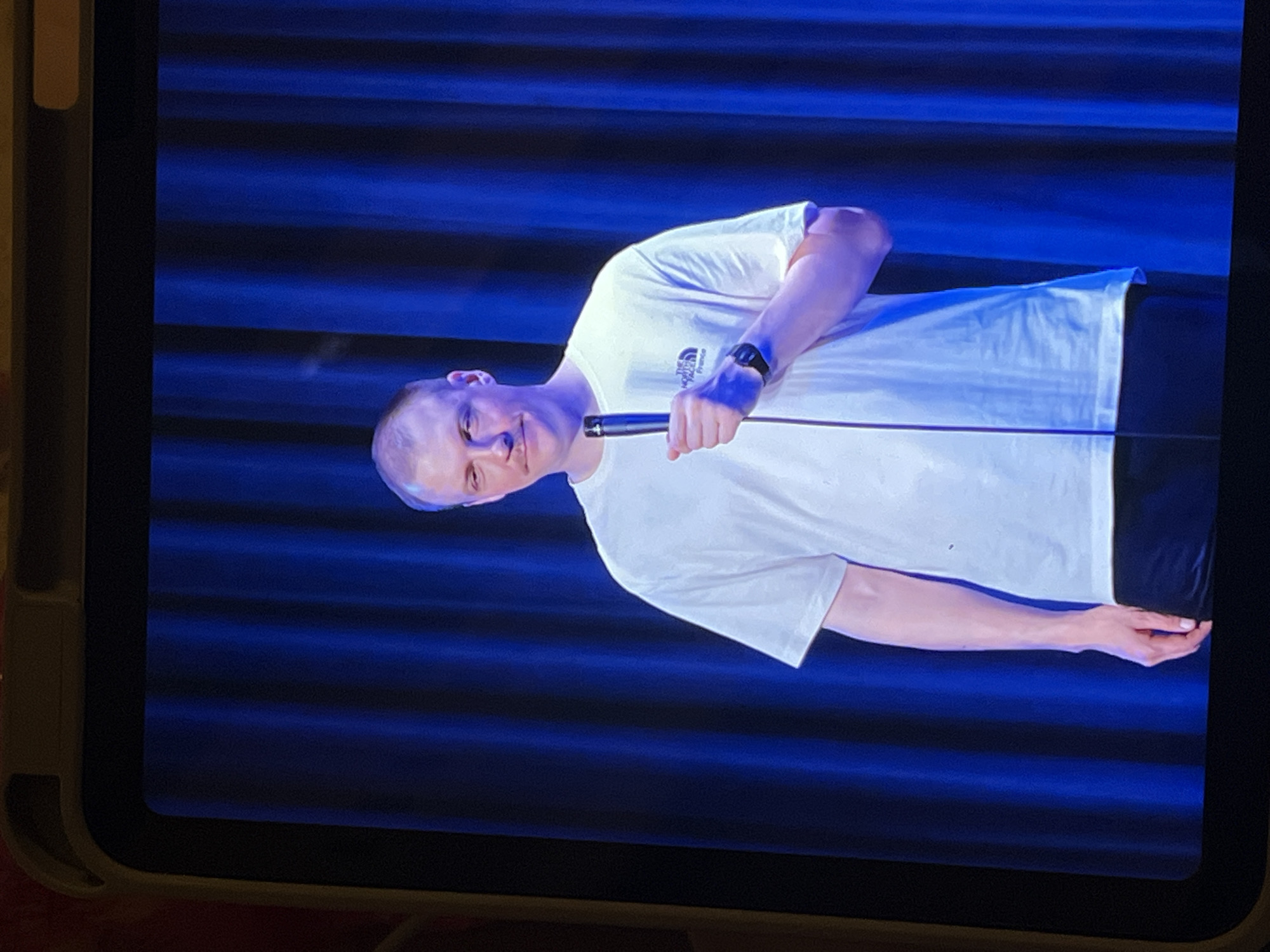 A man wearing a white t-shirt and black pants stands on a stage, holding a microphone. He has short, light-colored hair and a mustache. A blue curtain serves as the backdrop for the scene. The image is framed by a yellow border, suggesting it is a picture taken from a screen, likely a tablet or phone. There are no visible landmarks or context to place this scene in Genemuiden, Netherlands.