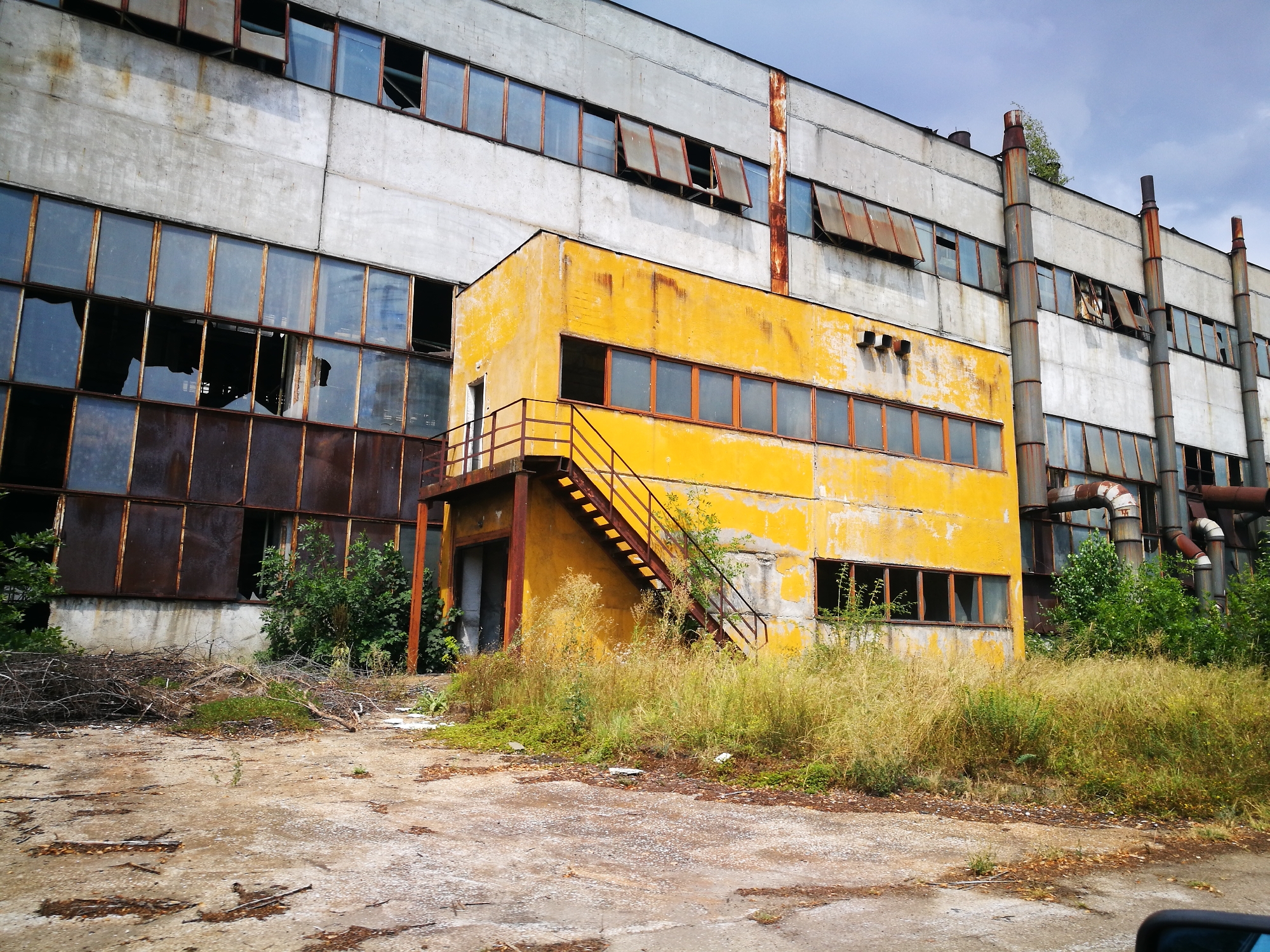 A wide, eye-level, outdoor shot shows an abandoned industrial building with a bright yellow annex on the left. The main building is a long, multi-story structure made of concrete, with many windows. Some of the windows are broken or missing, and some have rusted frames. The yellow annex is smaller and has a similar window style. An external metal staircase with a railing leads up to a door on the yellow annex. The area around the building is overgrown with tall grass and weeds, and there are piles of debris and branches on the ground in the foreground. The sky is overcast and cloudy. The scene depicts an area in a state of disrepair and neglect.