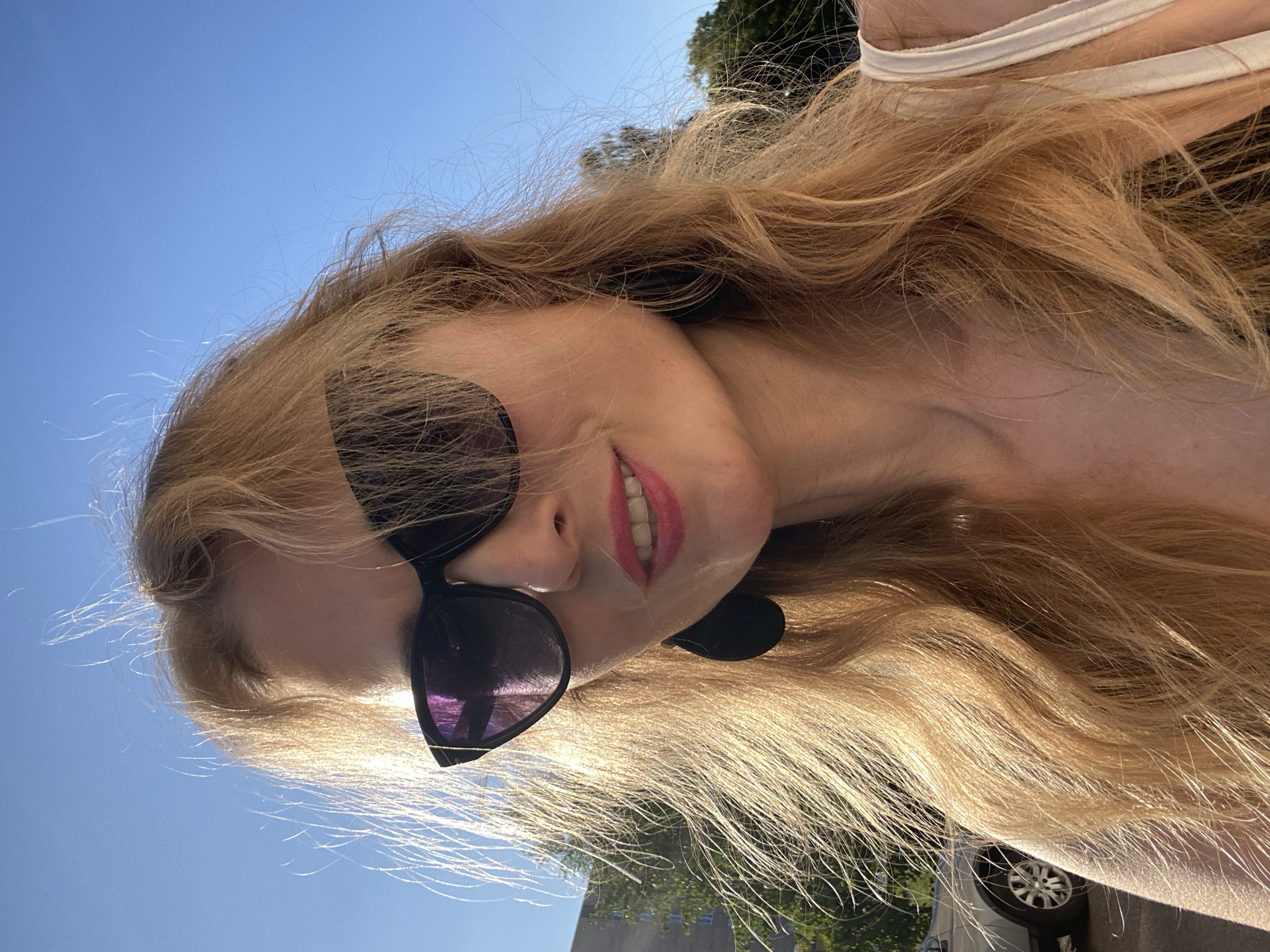 In a frontal, close-up selfie, a fair-skinned woman with blonde hair is captured outdoors against a clear blue sky. She wears large, black, cat-eye sunglasses that obscure her eyes. Her lips are painted a vibrant pink, and she has a slight smile. She is wearing a light-colored top and large, dark, teardrop-shaped earrings. Her blonde hair, with a reddish-blonde hue, is windswept and falls around her face and shoulders. In the background, a multi-story building and the tops of green trees are visible. A silver car is parked on the left side of the frame. The overall lighting suggests it is a sunny day, likely late afternoon due to the warm glow on her hair. The specific location is Lida, Belarus.