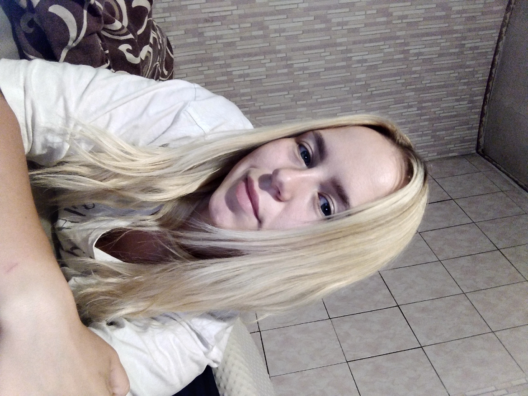 An upper body, medium shot, selfie-style photograph shows a young Caucasian woman with long, blonde hair, fair skin, and blue eyes. She is wearing a white t-shirt and appears to be lying down in bed. The background consists of tiled walls, one with a vertical striped pattern and the other with a grid pattern. A patterned blanket is visible to the left of the woman, and a pillow is to her right. The lighting is dim and appears to be artificial. The woman is looking at the camera with a slight smile.