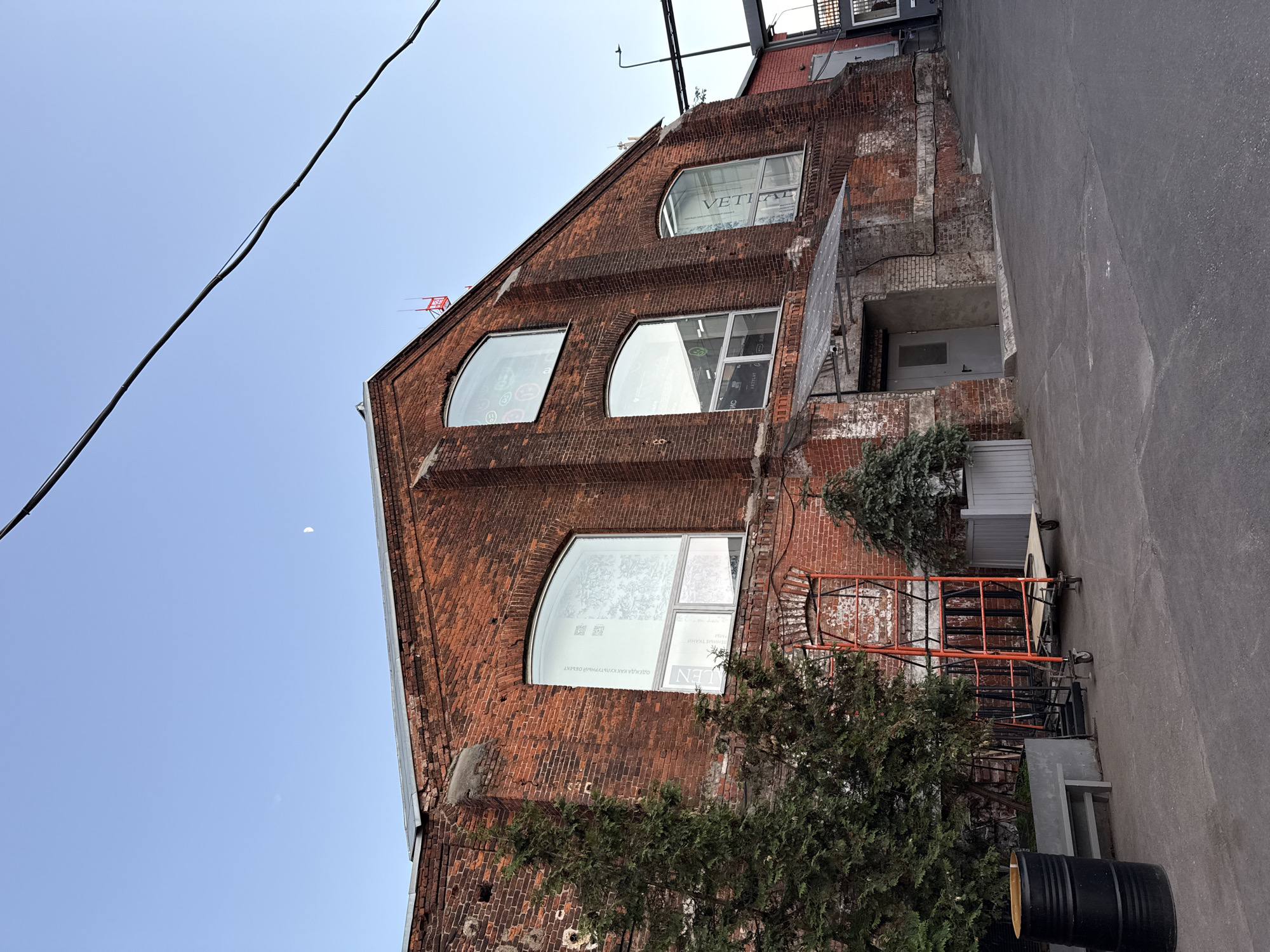 The image shows a three-story red brick building in Moscow, Russia. The building has three arched windows on each floor, and the top window has the word "VERTICAL" printed in the window. There are trees and shrubs partially obscuring the ground floor, and a metal scaffold is visible on the side of the building. There is a sidewalk and a street adjacent to the building. A power line is in the top left corner of the image. A quarter moon is in the sky.
