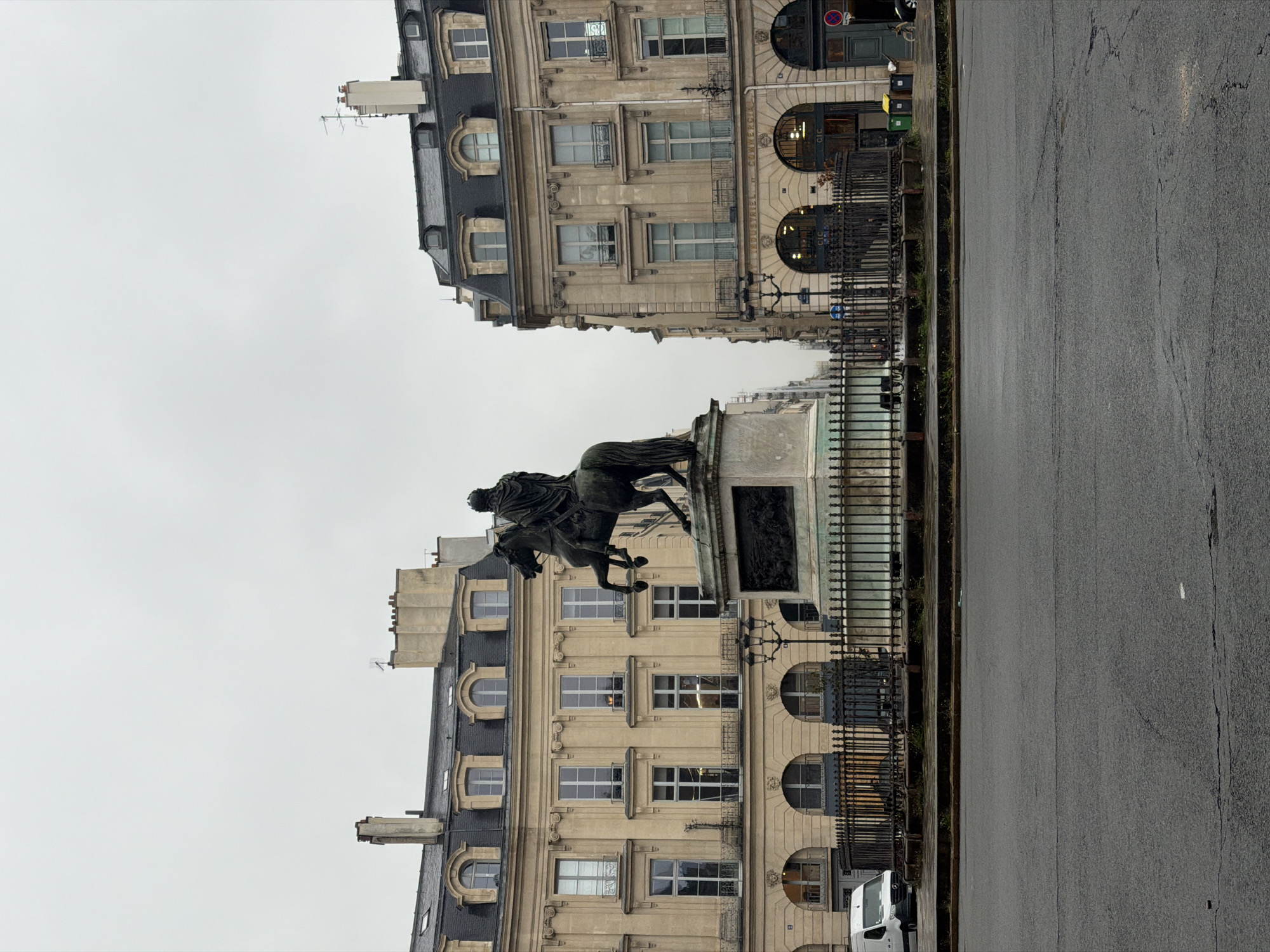 The image captures a street scene in Paris, France. The focal point is an equestrian statue situated on a stone pedestal, enclosed by a wrought-iron fence. Architectural buildings flank the street, showcasing a consistent Parisian style with multi-story facades, symmetrical window arrangements, and dark-sloped roofs with dormers. The street pavement appears wet, possibly from recent rain, and stretches along the right side of the frame. A white van is parked in the lower right corner of the image. The sky is overcast, lending a muted tone to the scene. The statue and buildings are positioned in a way that suggests they are located in a prominent square or public space.