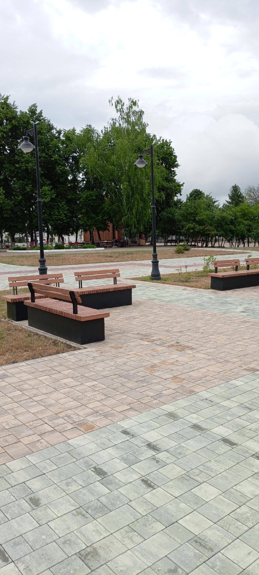 The image depicts an outdoor public area, likely a park or pedestrian zone in the city of Kirsanov, Russia. In the foreground, there are several modern wooden benches with black bases arranged on a paved pathway. The pathway is composed of light-colored rectangular tiles in a repeating geometric pattern, with sections of different colors, suggesting a designed walkway.
Behind the benches, a line of trees forms a backdrop, with a more densely wooded area further in the distance. Two ornate black lampposts with vintage-style lamps stand tall, one closer to the foreground and another further back. The sky above is overcast, with a muted, grey cloud cover.
In the middle ground, to the left, a wooden structure, possibly a pavilion or gazebo, is visible. Several more benches are situated around this structure. A few indistinct figures can be seen seated on these benches, suggesting the presence of other park visitors. The ground between the paved areas and the trees is covered in grass and dry foliage. The overall impression is one of a quiet, functional urban green space on a cloudy day.