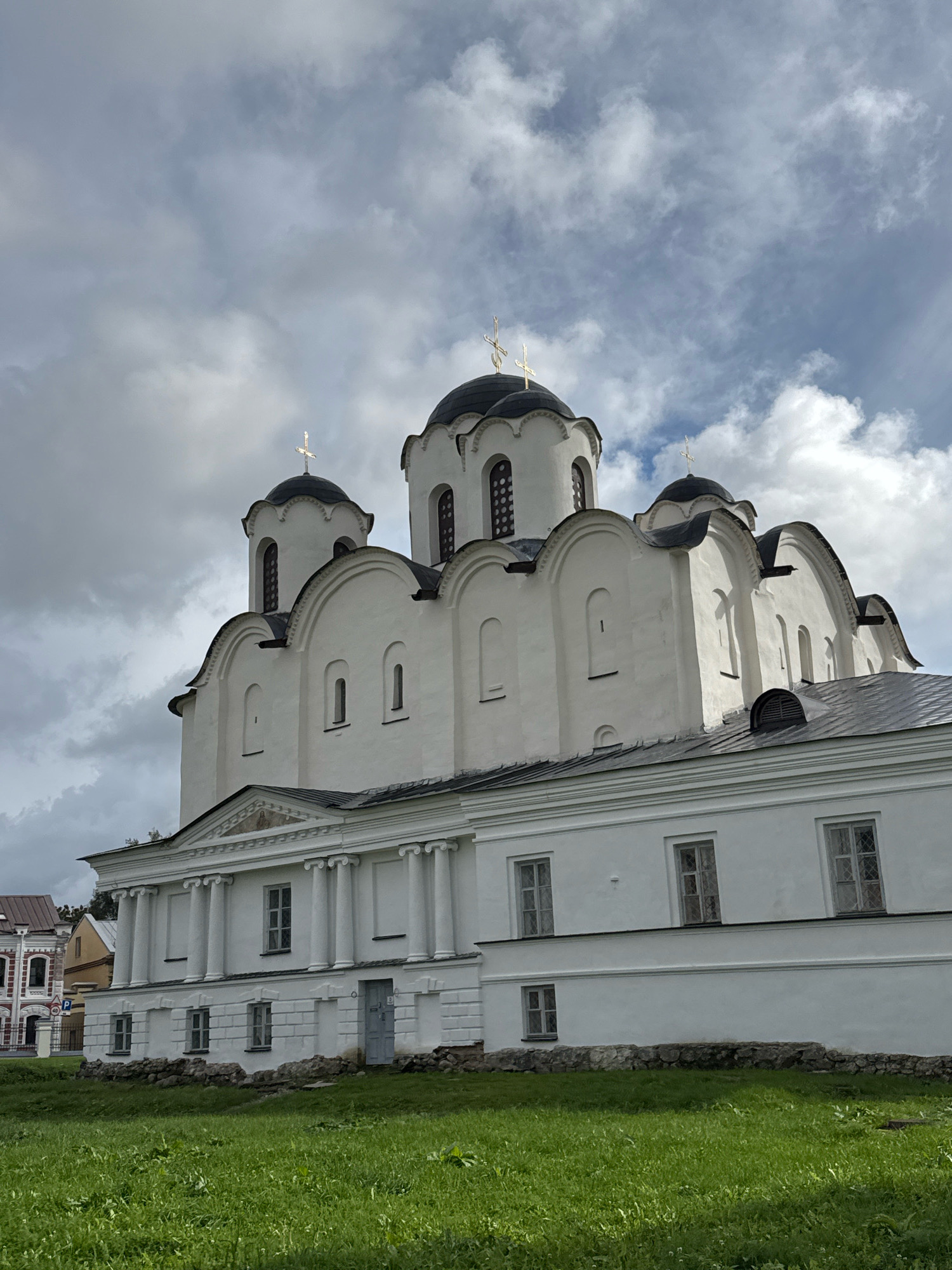 This is a photograph of the Saint Sophia Cathedral, a prominent landmark in Veliky Novgorod, Russia. The white cathedral features multiple dark domes topped with gold crosses, indicative of Orthodox Christian architecture. The building exhibits arched windows and a classical portico with columns on its lower level. The surrounding area is a grassy field, and the sky is partly cloudy with a mix of gray and white formations. In the background, to the left of the main structure, a portion of another building with red and white brickwork is visible.