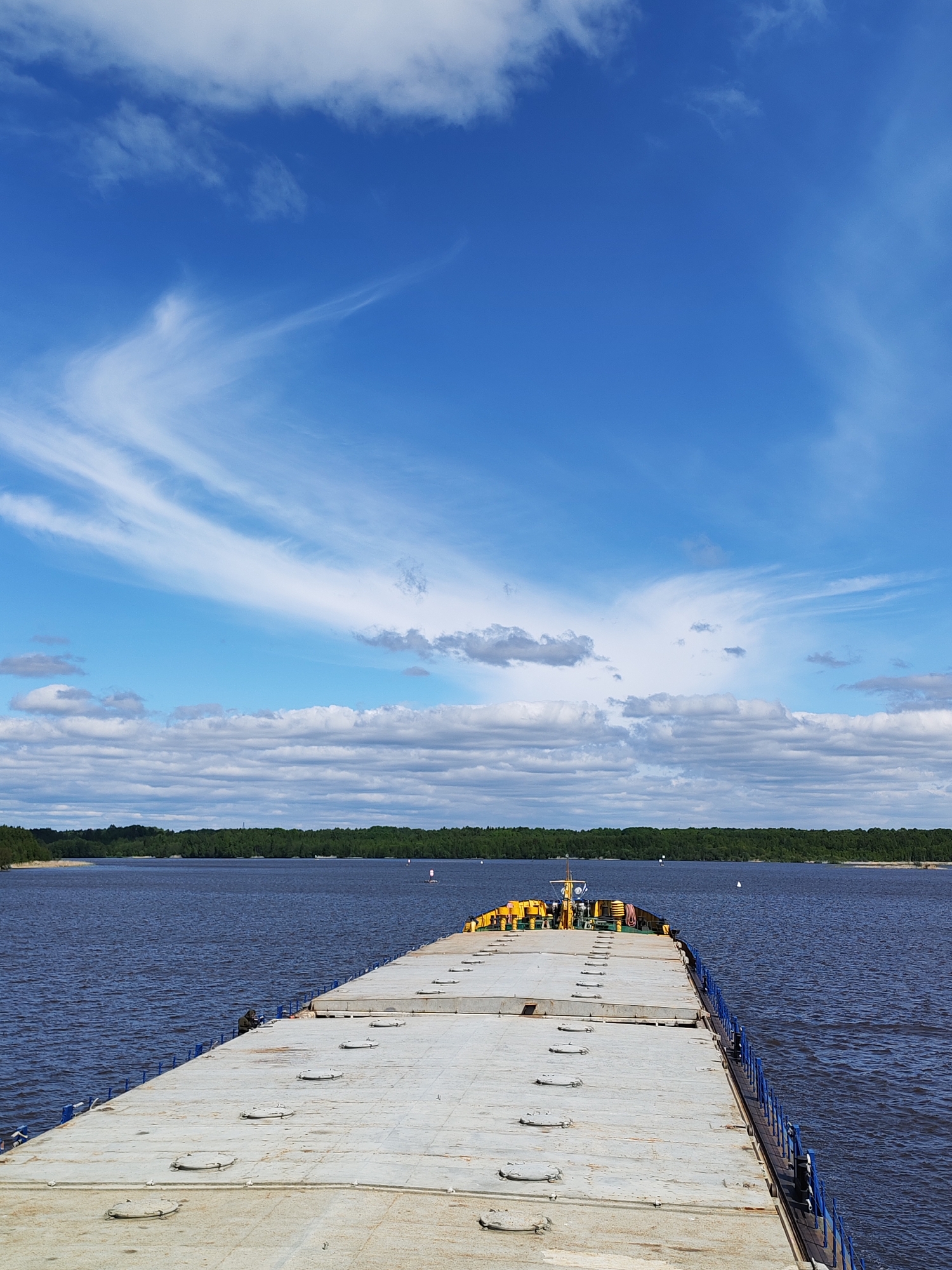 The image shows a view from the deck of a barge or large raft on a body of water. The deck is grey and features several circular access hatches. A low blue railing lines the edge of the deck. In the distance, a line of trees is visible along the far shore. The sky is blue with wispy cirrus clouds above and a layer of cumulus clouds closer to the horizon. The water is dark blue. There is no evidence to confirm the location is in the city of Novodevich'e, Russia.
