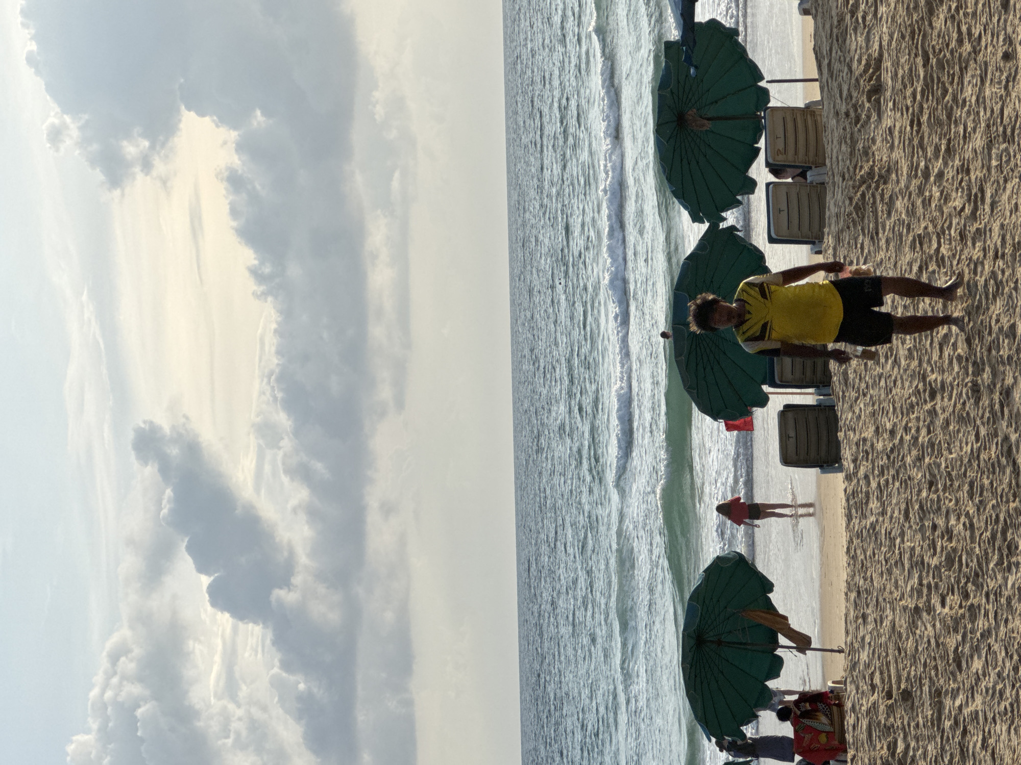 In Karon, Thailand, a sandy beach scene unfolds under a cloudy sky. The ocean, with its gentle waves, stretches to the horizon. In the foreground, beach umbrellas provide shade. A man wearing a yellow shirt and black shorts walks on the sand. In the background, several people are visible, some near the water's edge and others relaxing under umbrellas or on lounge chairs. The overall atmosphere is that of a typical day at a beach resort.