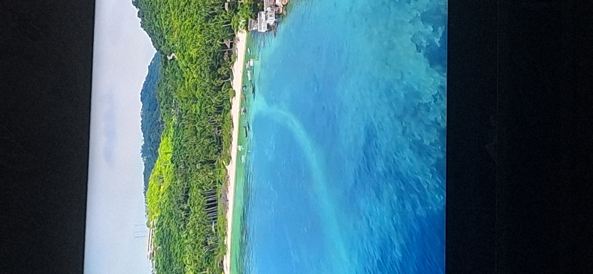 The image shows a tropical coastal scene. In the foreground, the clear blue ocean water exhibits a light-colored streak, possibly indicative of a sandbar or a change in water depth. Scattered across the water are several small boats. Along the shoreline, a white sand beach is lined with numerous palm trees. Behind the beach, lush green hills rise, covered in dense vegetation and trees. A winding road can be seen ascending one of the hills. On the top of one of the hills, a large building complex is visible, with several tall, slender structures resembling antennae or pylons. On the far right of the image, a small cluster of buildings is situated on rocky outcroppings near the water. The sky above is overcast and pale. The image is a still photograph, likely taken from an elevated viewpoint. There is no indication of the city of Bila Tserkva, Ukraine, in this image, as it depicts a tropical environment inconsistent with Ukraine's geography.