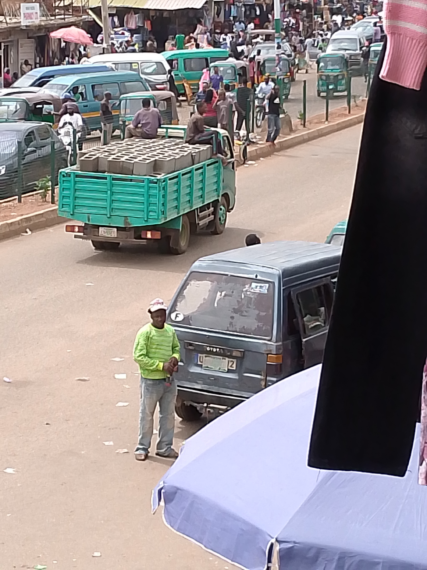 The image shows a busy street scene in an urban area. A green truck loaded with gray concrete blocks is driving away. A man in a striped green shirt, gray pants, and a baseball cap stands next to a gray Toyota van with its door open. In the background, there are many pedestrians and vehicles, including green and blue vans, and "keke" tricycle taxis. There's an array of shops with awnings visible in the background. The location is potentially a market or commercial district. Foreground elements obscure the view, including a blue umbrella and hanging clothes.