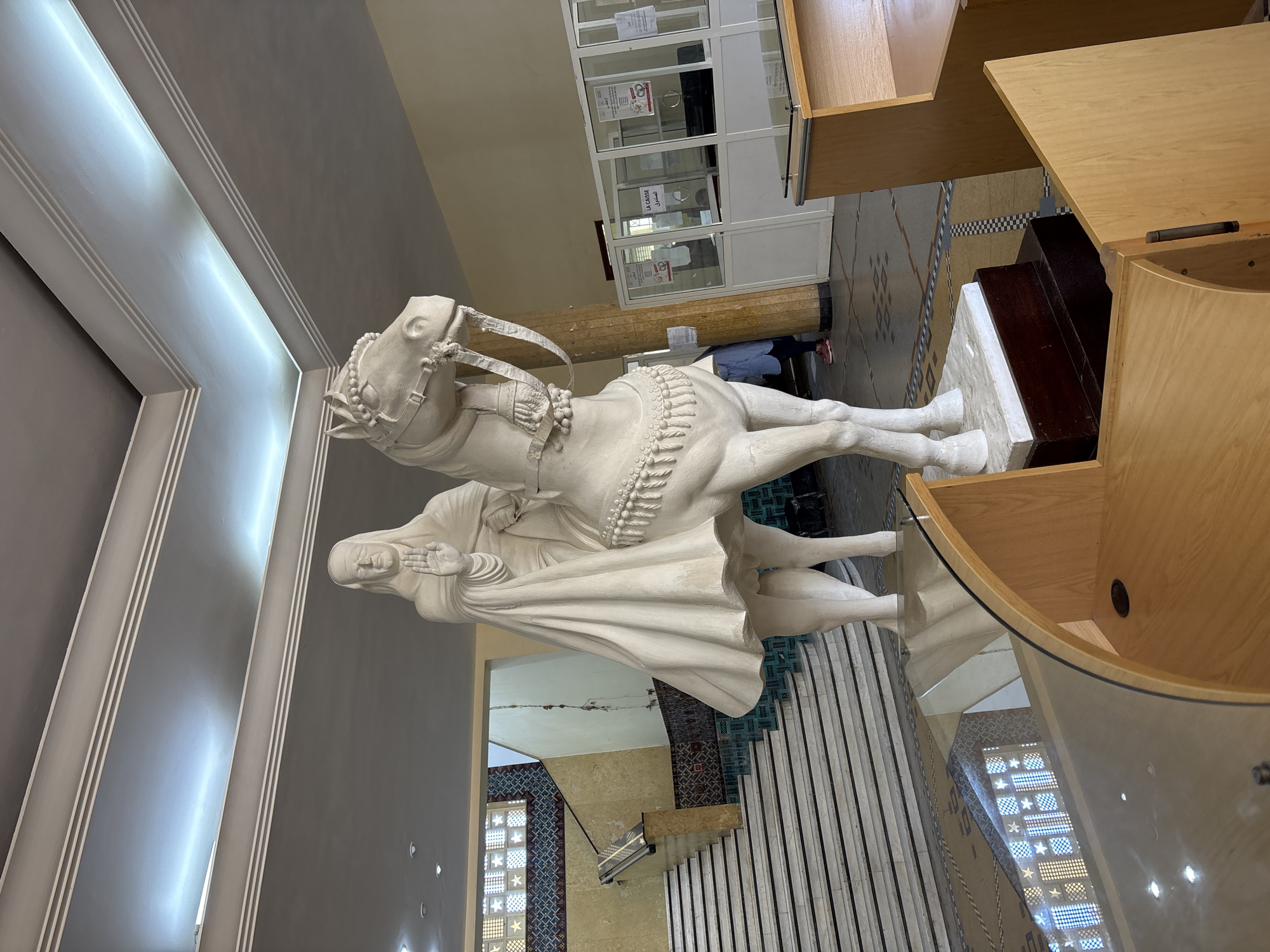In an interior space with a grey ceiling featuring recessed linear lighting and tiered trim, a large white sculpture of a person on a horse is prominently displayed. The figure, draped in flowing robes and wearing a head covering, has its right hand raised in a gesture of blessing or greeting. The horse is adorned with decorative bridle and breastplate. To the left of the sculpture, a wide staircase with white marble steps and dark treads ascends. The staircase area is decorated with patterned tiles in blue, gold, and red, and features a window with star-shaped panes. In the background, to the right of the sculpture, is a reception area with glass partitions. Signs are visible on the glass partitions, one reading "LA CAISSE" in French. Two individuals are partially visible behind the counter. In the foreground, there are wooden furniture pieces with glass surfaces, obscuring the lower portion of the sculpture and the floor. The floor itself is tiled with a geometric pattern.