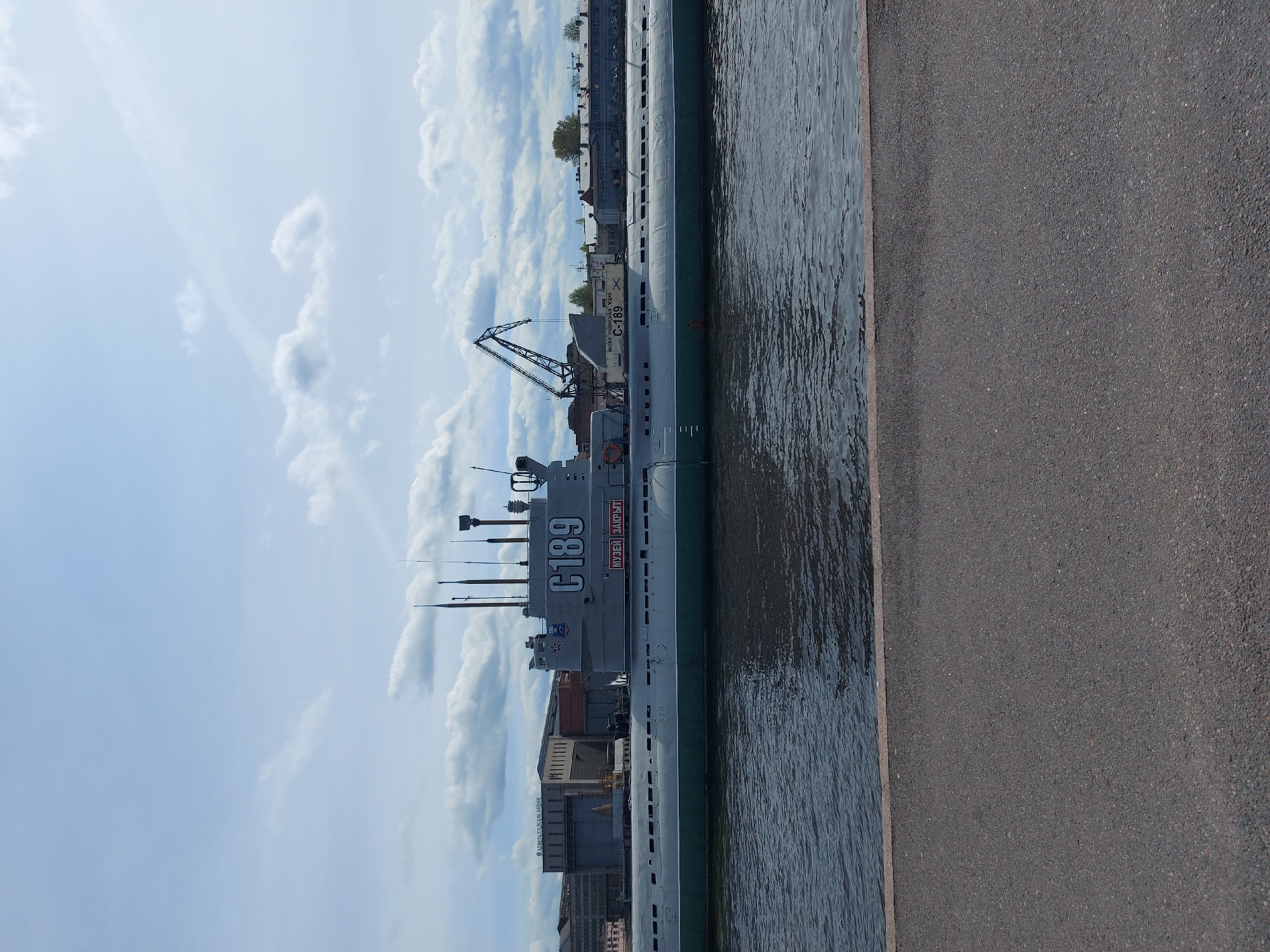 The image shows a Soviet submarine, specifically the C-189, docked on the Neva River in Saint Petersburg, Russia. The submarine, now a museum ship, is prominently labeled with "C189" and Russian text. It sits alongside a concrete walkway, with the river extending away from the vessel. The background features various buildings and industrial structures, as well as a crane on a pier. The sky is partially cloudy.