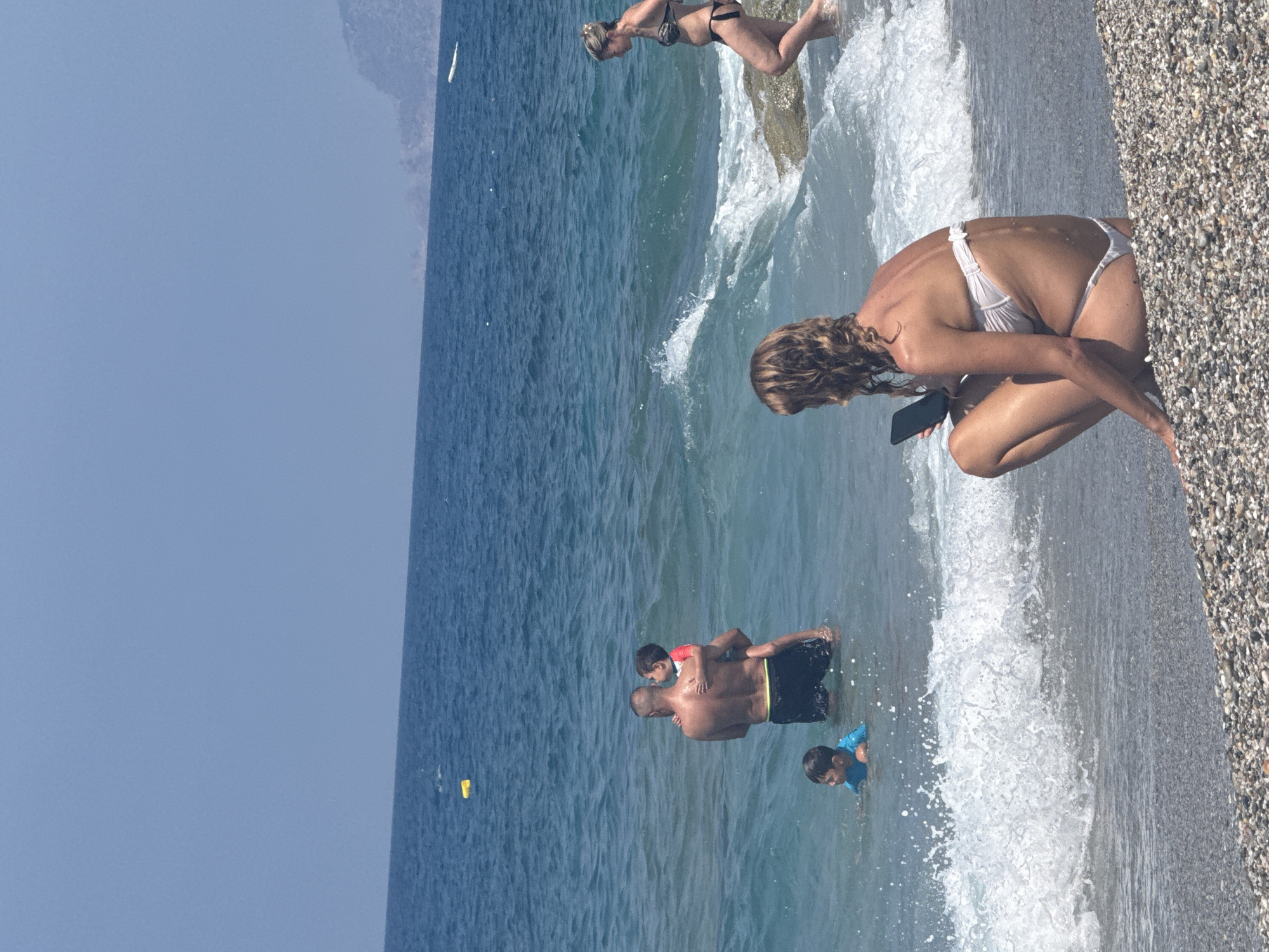 This image depicts a beach scene in Alanya, Turkey. In the foreground, a woman wearing a white bikini is crouching on the pebble beach, looking at her phone. Behind her, the ocean waves are breaking. Further out in the water, a man is holding a young child, while another child swims nearby. In the background, another woman in a bikini is walking in the water near some rocks. The water is a deep blue, and the sky is clear and light blue. Mountains are visible in the distance on the right side of the image. A yellow object, possibly a buoy, is floating in the water in the upper left quadrant of the image. The overall scene suggests a sunny day at a tourist beach.