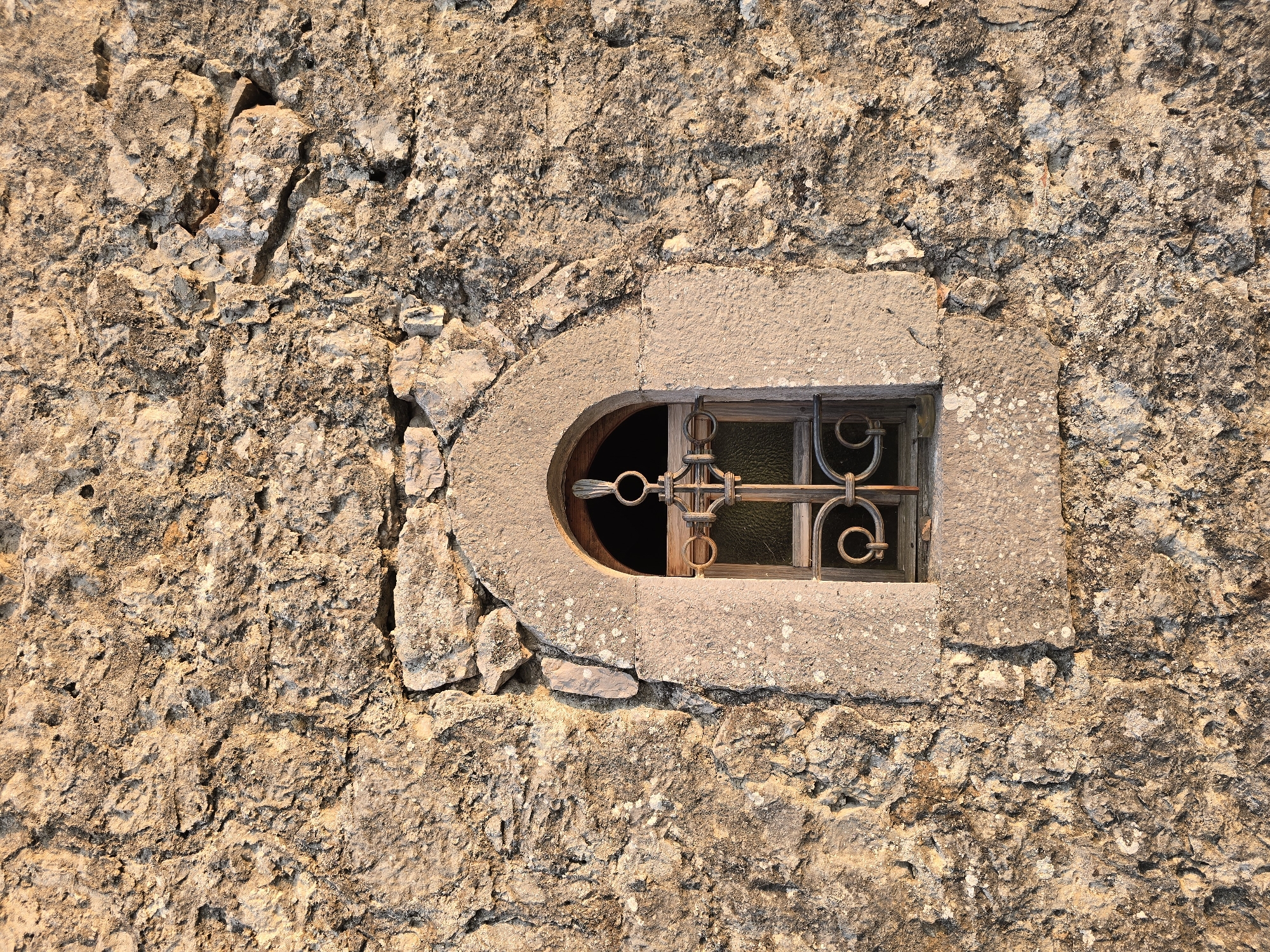 The image displays a close-up, eye-level view of a small, arched window set into a rough, textured stone wall in Medun, Montenegro. The wall is constructed from irregularly shaped, light-colored stones, some of which are large and prominently featured, while others are smaller and fill the gaps. The stone surface exhibits a rough, weathered texture, with visible cracks, pits, and patches of lichen or moss. The window itself has a stone frame with a rounded top. Within the frame, a wooden-paned window is visible, covered by a dark metal grate. The grate features an ornate design with scrolled elements and a central vertical bar. The light appears to be warm, suggesting it might be late afternoon or early morning, casting subtle shadows and highlighting the uneven surfaces of the stone. No people are visible in the image. The overall impression is one of age and historical resilience.