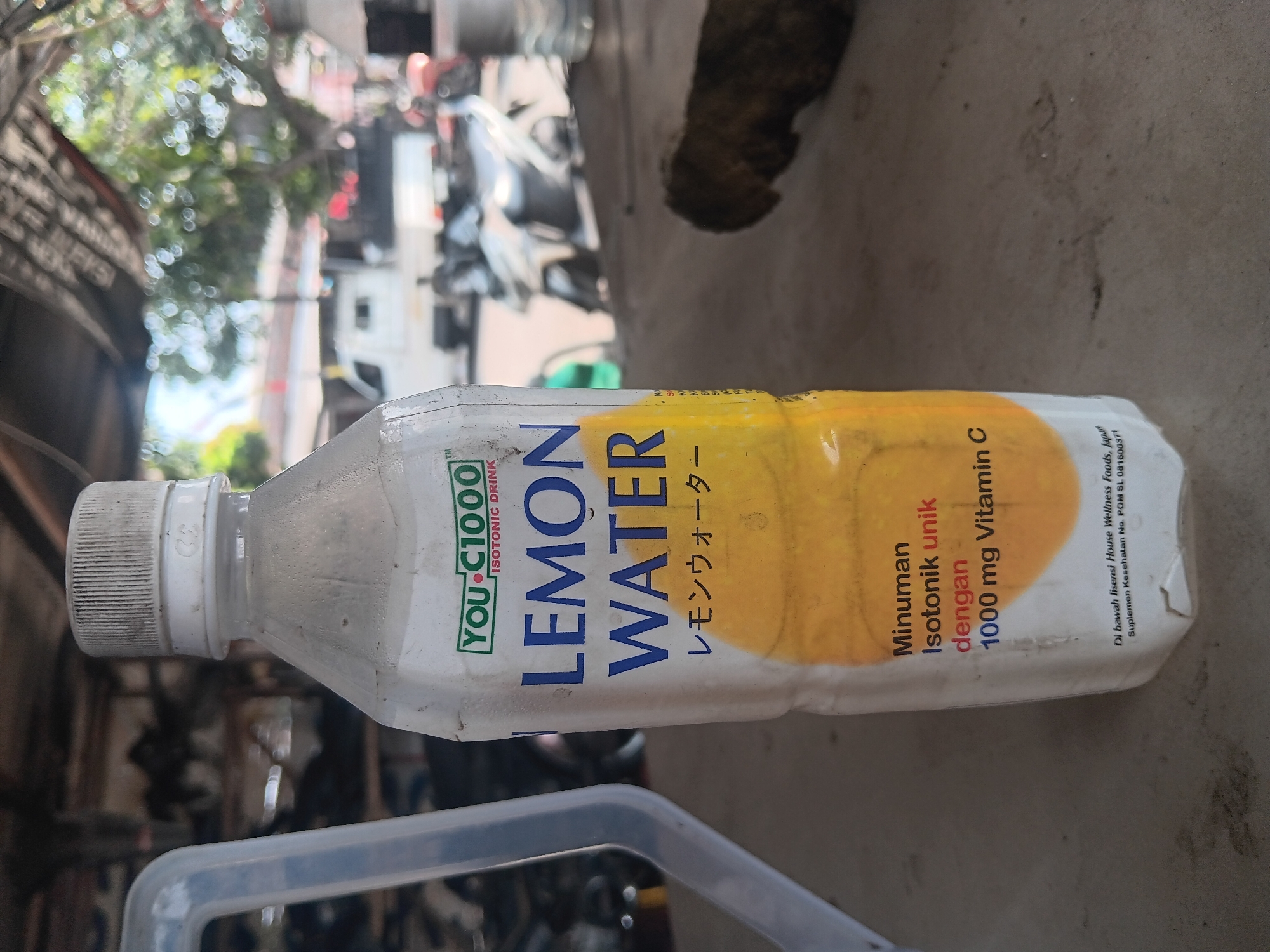 The image displays a white plastic bottle of "YOU-C1000 LEMON WATER" with Japanese text below. The bottle is sitting on a grey, textured surface, likely a table or counter. The background is blurred but suggests an outdoor or semi-outdoor setting with trees, buildings, and several motorcycles. There is a suggestion of a market or roadside stall. The scene appears to be in a warm, possibly tropical environment, indicated by the bright sunlight and foliage. The bottle itself shows some condensation and dust, implying it has been exposed to the elements. There is no direct human presence visible in the immediate vicinity of the bottle, although blurred figures can be seen in the background, suggesting activity. The overall impression is that of an everyday scene in a public space in Tlogo, Indonesia, likely a place where beverages are sold or consumed.