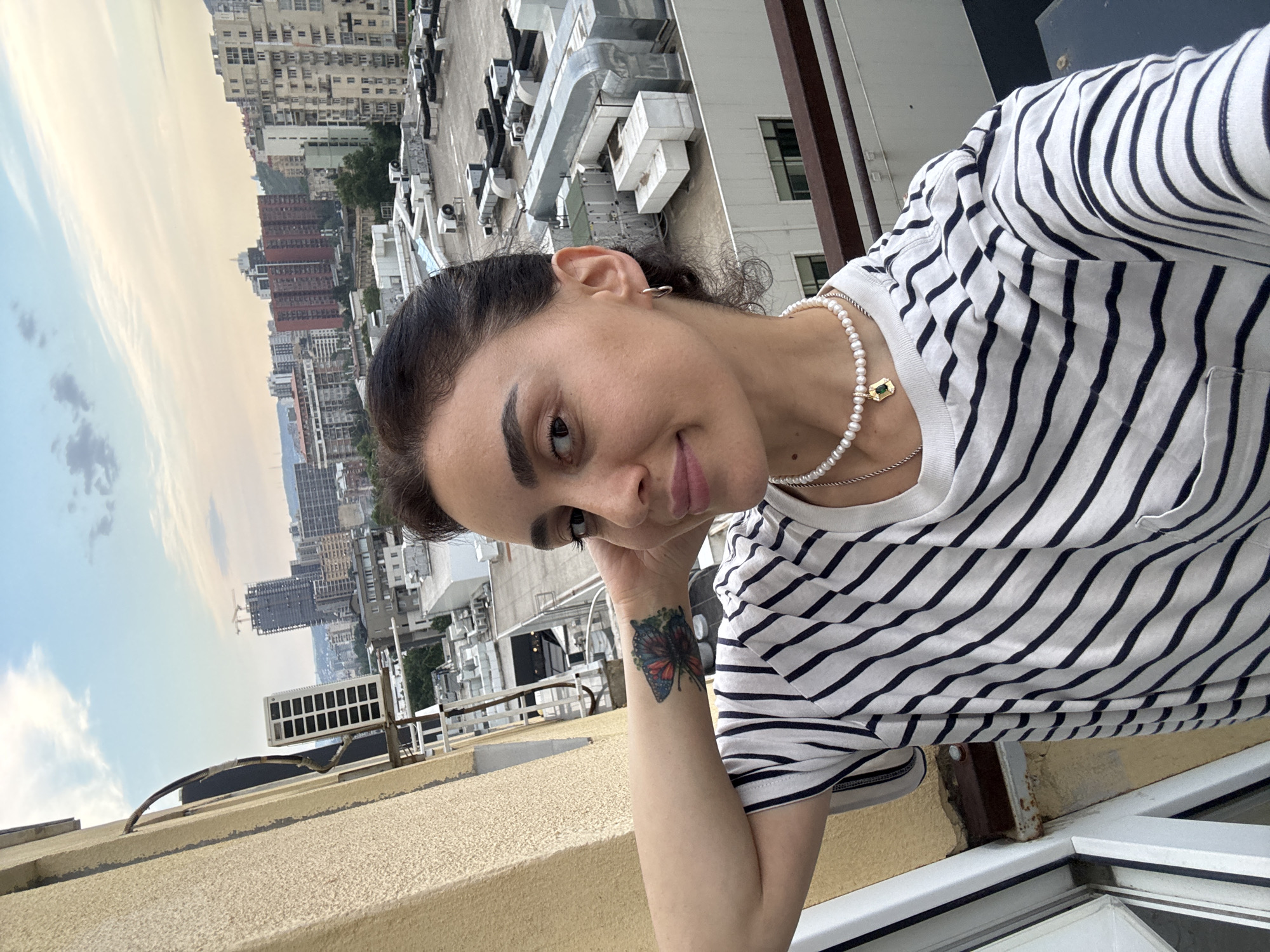 A selfie taken by a woman with dark hair pulled back, wearing a white t-shirt with black horizontal stripes and a pearl necklace with a green pendant, is captured on a balcony in Tbilisi, Georgia. She is looking towards the camera with a slight smile, her left arm resting on her head, revealing a butterfly tattoo on her wrist. In the background, the cityscape of Tbilisi unfolds, with buildings of various heights and architectural styles, construction cranes, and an overcast sky. The balcony railing is visible in the foreground, and the textured wall of an adjacent building is to the left. The scene depicts a moment of leisure with an urban vista.