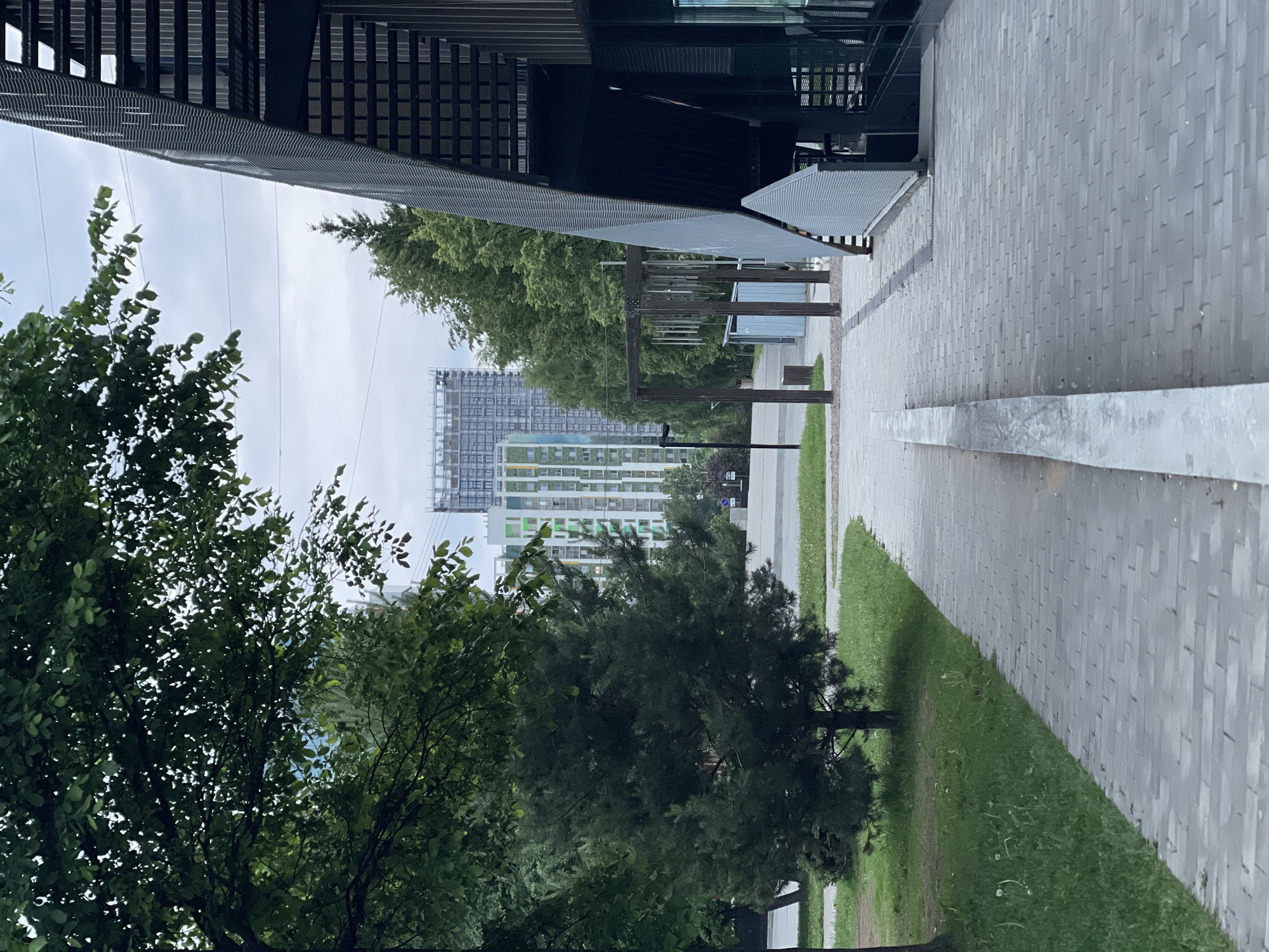 This image depicts an outdoor urban or suburban environment in the city of Unknown, Unknown, characterized by a paved pedestrian pathway, green spaces, and architectural structures under an overcast sky.
In the immediate foreground, a light grey interlocking brick pathway extends from the bottom center into the distance, slightly curving to the left. A raised concrete curb or separator runs along the left edge of this pathway. To the left of the pathway is a strip of vibrant green grass, featuring a young, dark green coniferous tree with a visible trunk and branches. Further left, partially obscured, is the trunk of another deciduous tree. To the right of the pathway, the angular, metallic-grey facade of a modern building dominates the right side of the frame. This facade is composed of horizontal ribbed panels and possibly perforated sections, creating a textured, almost folded appearance. A dark, recessed opening suggests an entrance or sheltered area.
In the midground, the paved pathway continues, flanked by more green grass and mature deciduous trees with dense foliage on the left. On the right side of the pathway, a series of dark wooden posts are arranged, supporting a horizontal beam from which several metallic, tubular objects (resembling wind chimes) are suspended. Behind these posts, two small, light-colored utility sheds or enclosures are visible. A black lamppost stands upright on the left side of the pathway.
The background features additional trees and two prominent multi-story buildings. The closer building is a tall, contemporary residential structure with a light-colored (possibly white or light grey) facade, accented by vertical panels or balcony elements in bright green, yellow, and light blue. Behind and slightly to the right of this residential building, a taller, darker grey building appears to be under construction, with visible scaffolding or unfinished structural elements at its upper levels. The sky is uniformly overcast, exhibiting a light grey tone, suggesting a cloudy day without direct sunlight. There are no individuals, vehicles, or discernible activities beyond the implied construction in the distant background.