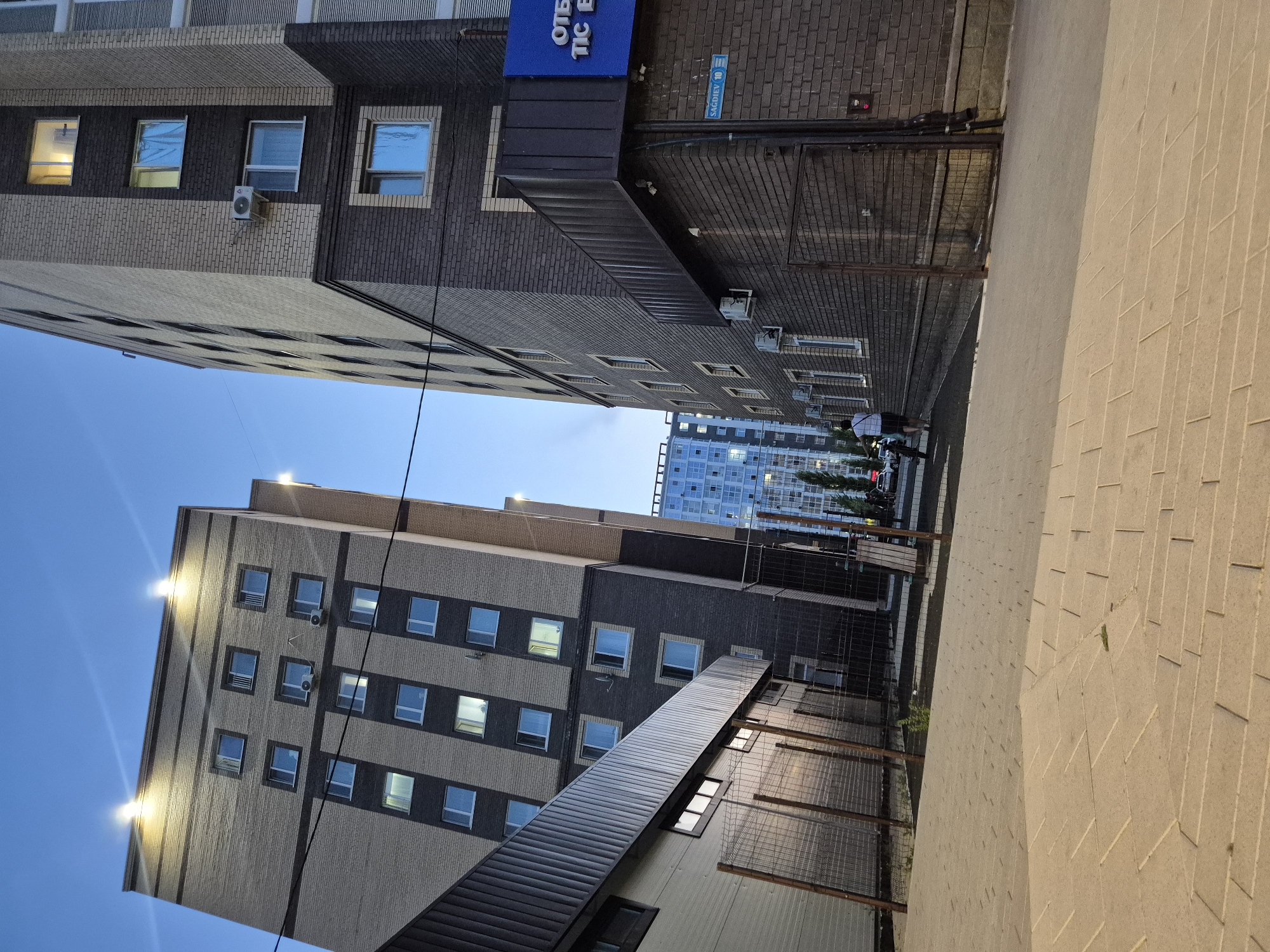 In Kokshetau, Kazakhstan, a low-angle, full-shot photograph captures a street scene at dusk. Two adjacent apartment buildings, constructed with a mix of beige and dark brown brickwork, dominate the frame. The buildings are illuminated by lights, highlighting their numerous windows. In the foreground, a paved walkway made of light-colored rectangular stones leads towards the buildings. A chain-link fence separates the walkway from a darker, lower structure on the left. Beyond this, the apartment buildings rise several stories. On the right side, a blue sign with white lettering is visible on one of the buildings, along with a street sign indicating "SAGDIYEV 10". A person is seen walking away from the camera towards the buildings, pushing a bicycle. The sky above is a clear, pale blue, suggesting early evening.