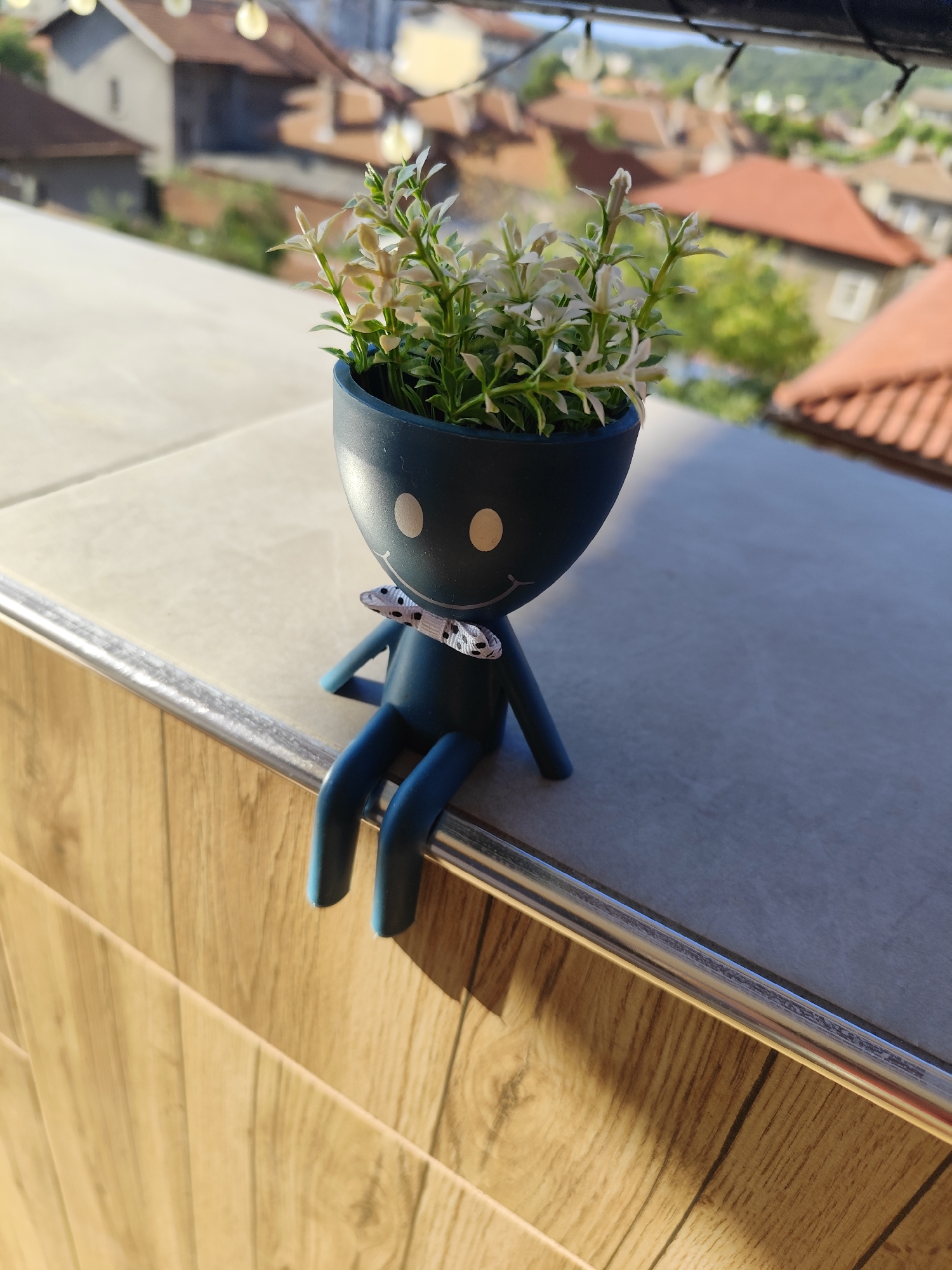 In Montana, Bulgaria, a small, dark blue ceramic pot shaped like a cartoon figure with a smiley face is perched on a tiled ledge. The figure has two white oval eyes and a small black and white polka-dot bow tie. It holds a sprig of artificial green and white plants in its head. The figure's blue legs dangle over the edge of the ledge, which overlooks a neighborhood with red-tiled roofs and scattered trees. String lights are visible in the background, suggesting an outdoor setting, possibly a balcony or windowsill. The scene is illuminated by natural sunlight, casting shadows on the wooden-patterned surface below the ledge.