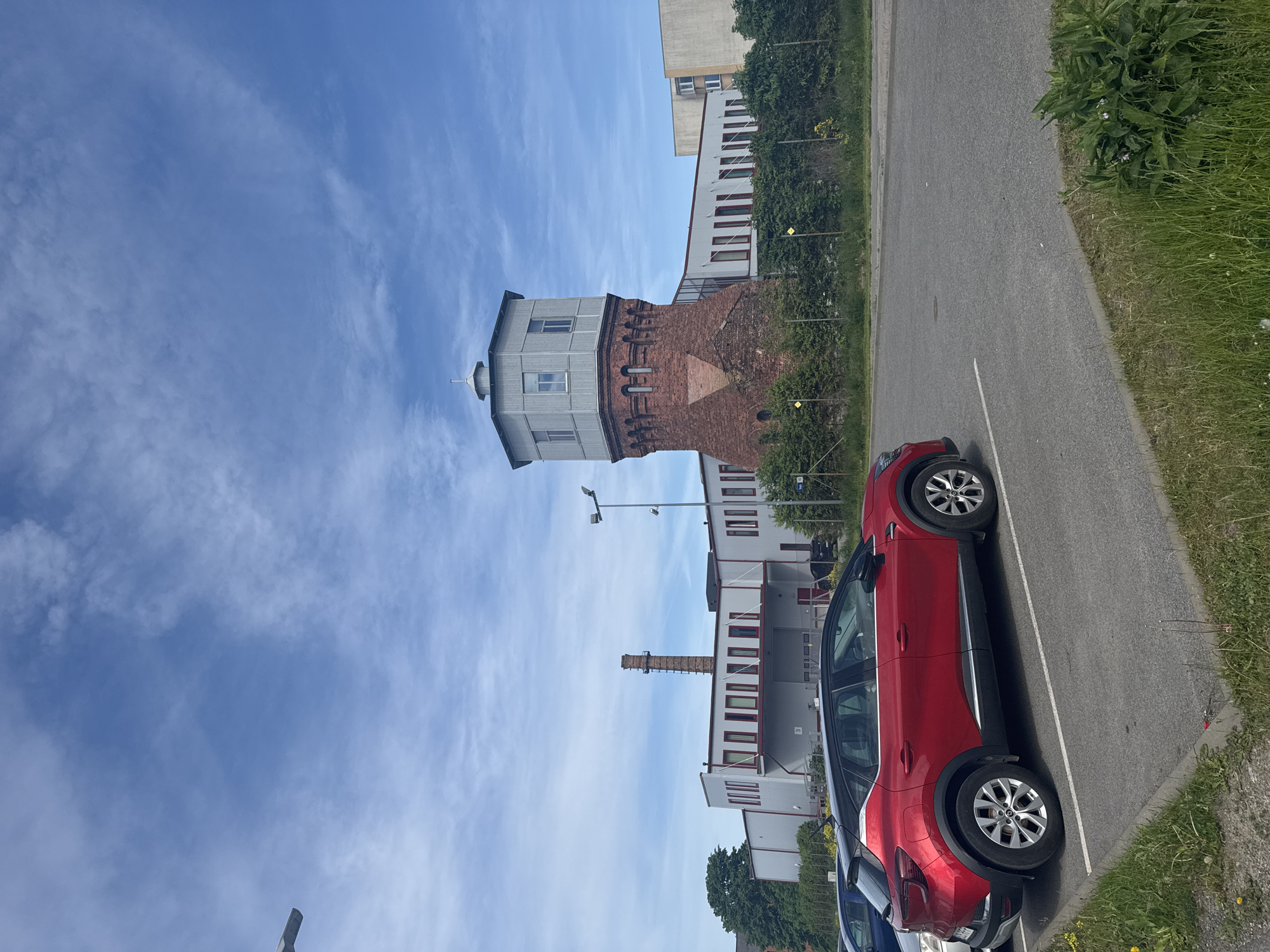 The image shows a red car parked in a lot, with a brick water tower and a modern industrial building in the background. The scene is set on a sunny day with a partially cloudy sky. The water tower, with its distinct brick base and gray, slatted upper section, stands to the right of the frame. Adjacent to it is a long, rectangular building with a light-colored facade and a series of windows. A tall, narrow brick chimney rises from the roof of this building on the left side. The parking lot is asphalt, with white lines marking the parking spaces. Green grass and some bushes are visible along the edge of the parking lot and surrounding the buildings. The water tower is located in Tallinn, Estonia.