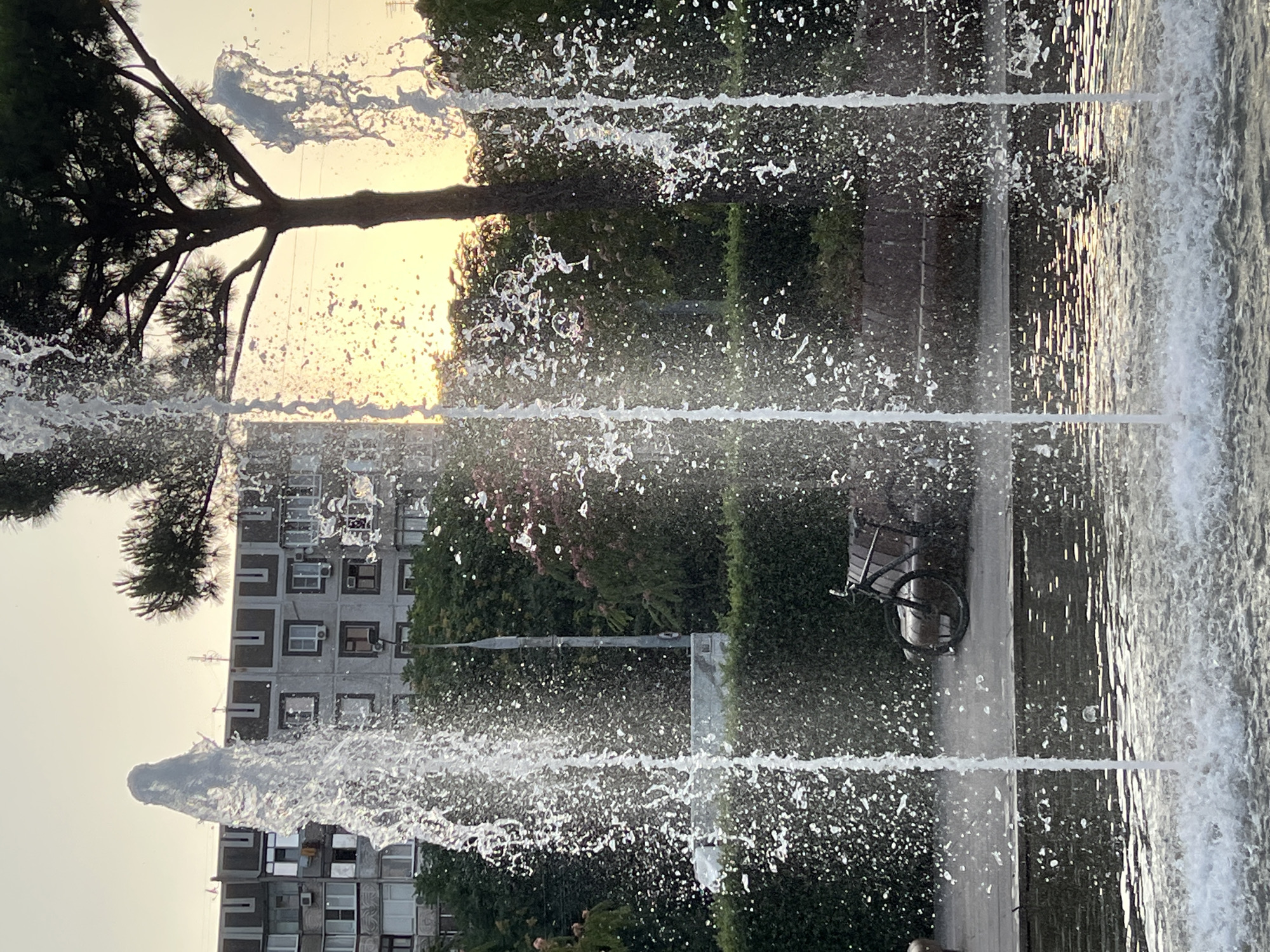 The image shows a fountain with water spraying in vertical streams. The scene is set outdoors, with a building, trees, and a pathway visible in the background. In the middle ground, a bicycle is parked near a bench. The sky is bright, suggesting it is daytime, possibly late afternoon or early evening due to the warm light. The overall mood is calm and serene, with the water from the fountain creating a sense of freshness. The location is identified as Krasnodar, Russia. There is no visible text in the image.