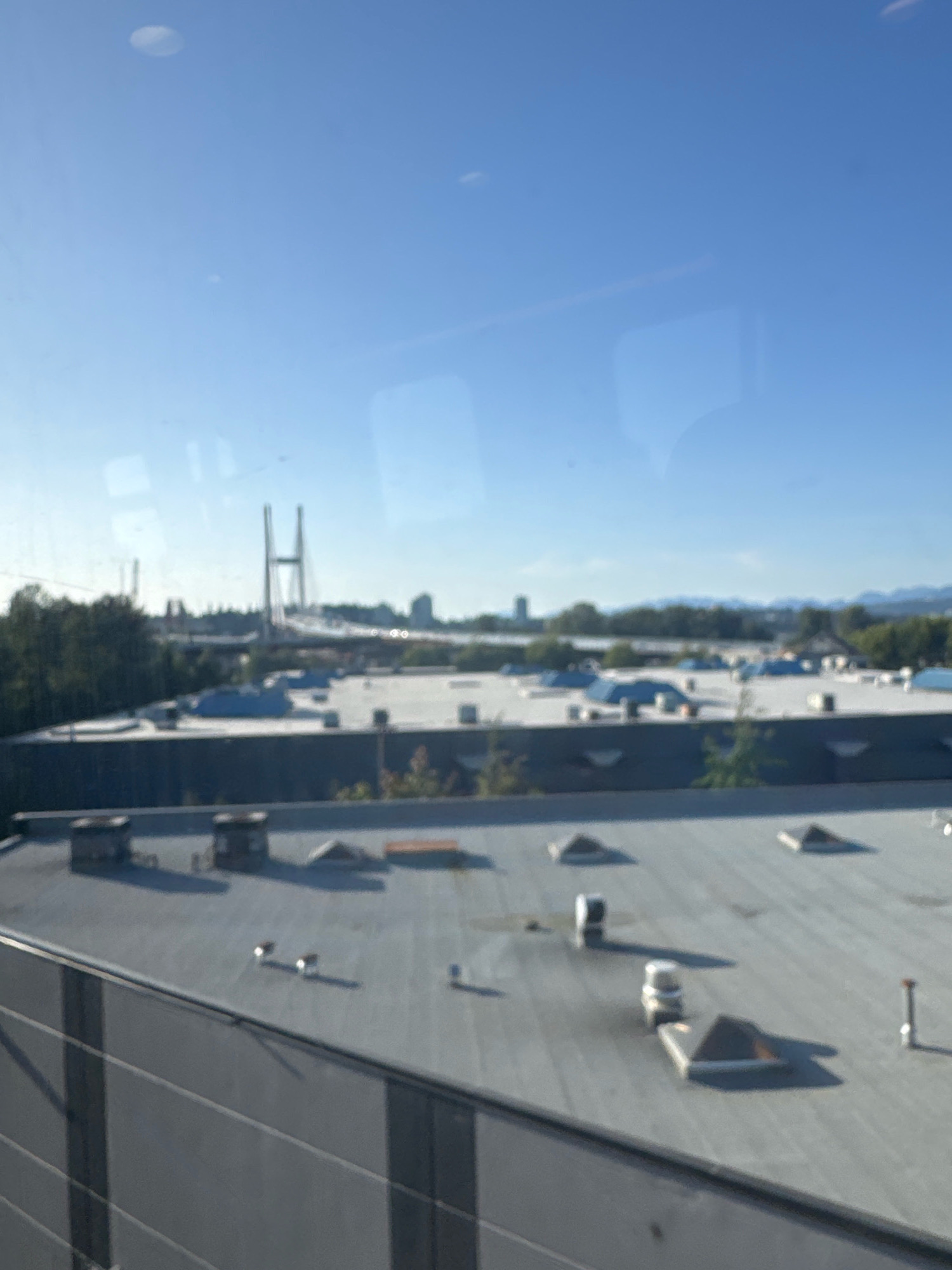 This is a daytime exterior photograph taken from an elevated vantage point, likely looking out from a building. The scene depicts a cityscape with industrial or commercial buildings and a bridge in the distance under a clear blue sky.
In the foreground, the flat, grey roof of a modern building is visible, featuring various mechanical units and rooftop structures such as vents, skylights, and what appear to be HVAC units. The roof is slightly wet in some areas. The side of another building with a grey, corrugated facade is also prominent in the lower portion of the frame.
Further into the scene, a long, flat-topped building with a white roof is present, also equipped with multiple rooftop units and some blue-covered sections, possibly for waterproofing or insulation. Beyond this building, a line of green trees forms a natural barrier, behind which a city skyline with several tall buildings is visible. The most striking element in the background is a large cable-stayed bridge with two prominent towers. The bridge spans across a body of water or a wide expanse, and its design suggests it is a significant piece of infrastructure. Mountains are visible in the far distance on the horizon.
The image appears to be taken from within or near the city of Surrey, Canada. The presence of a large cable-stayed bridge and the urban development in the background are consistent with locations in the Metro Vancouver area. The specific bridge visible is likely the Alex Fraser Bridge, which spans the Fraser River and connects Surrey to Richmond and Delta. The overall impression is of a clear, sunny day in an urban or suburban industrial area.