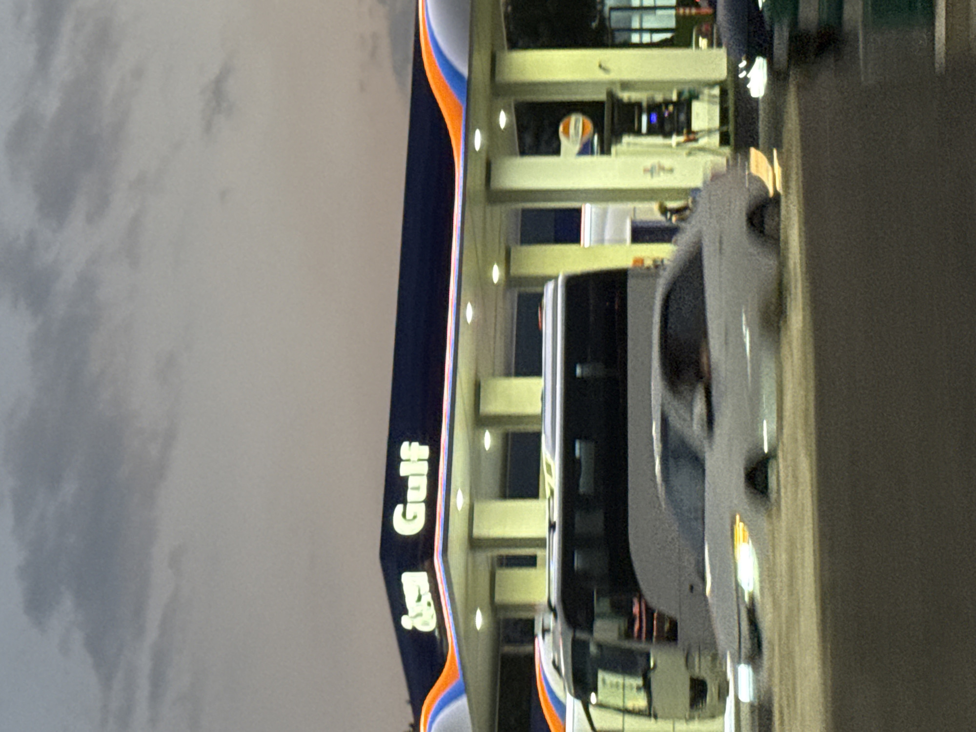 The image shows a Gulf gas station in Tbilisi, Georgia, with a blurred foreground of cars and a bus. The gas station sign prominently displays "Gulf" in white, illuminated lettering. The sky above is overcast with a muted twilight glow. The architecture of the gas station features a dark blue roof with colorful orange and blue accents. In the foreground, a white car is captured in motion, its headlights illuminated, suggesting it is driving past. A bus is also visible, partially obscured by the car. The overall scene depicts a typical urban environment at dusk, with the focus on the branded gas station.
