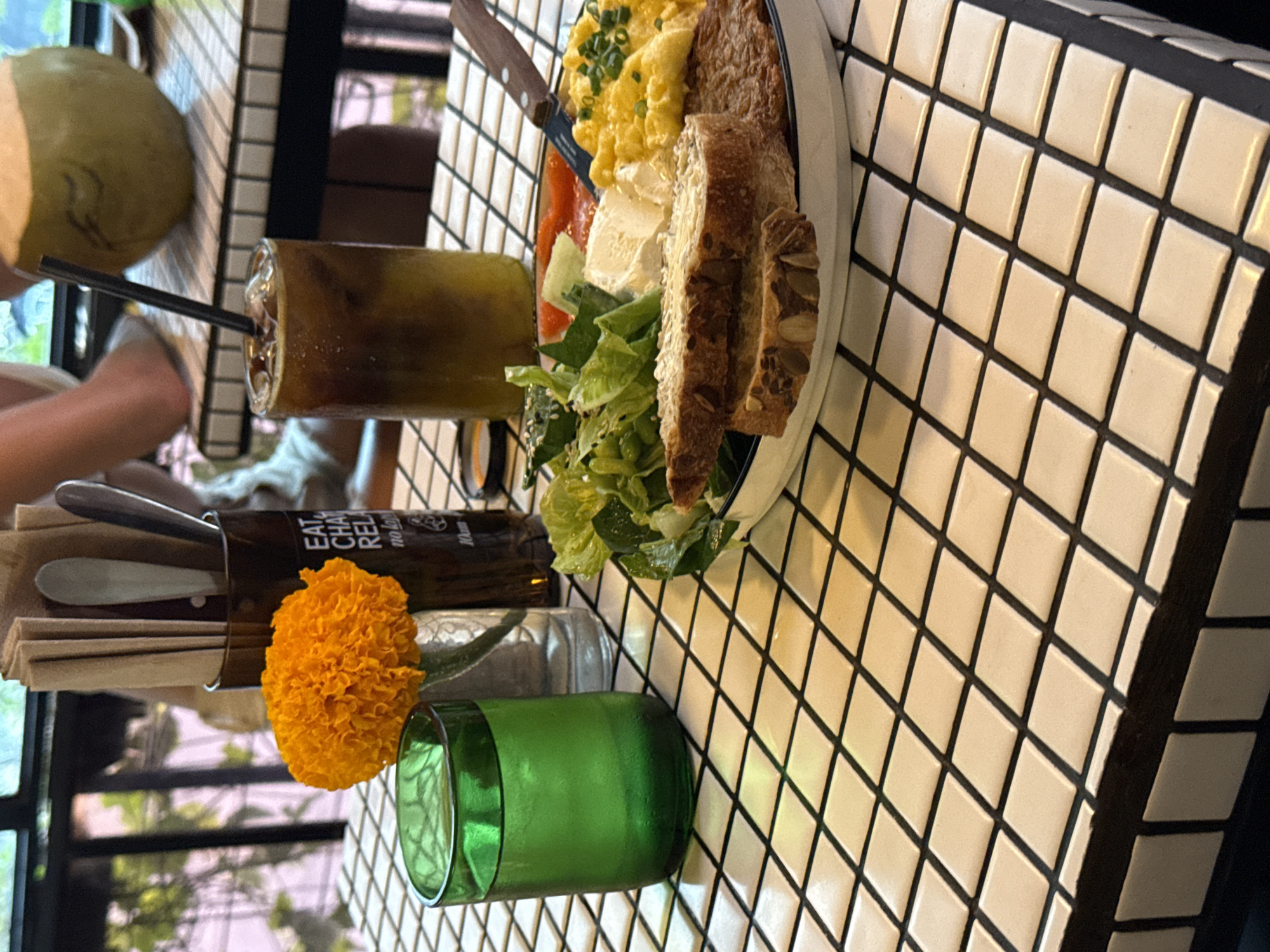 The image depicts a table setting in what appears to be a cafe or restaurant in North Kuta, Indonesia. The table has a black and white tiled surface, set with an arrangement of food and beverages. There is a plate with scrambled eggs, bread, salad, and other ingredients, likely a breakfast or brunch dish. Next to the plate, there's a tall glass filled with a layered drink, possibly coffee or tea. A coconut can also be seen on the table. A small green vase containing an orange flower is also visible near cutlery and napkins. Part of a person's arm is visible on the left of the image, suggesting someone is seated at the table. The scene is bathed in natural light, with a view of greenery outside the window, characteristic of the tropical environment of North Kuta, Indonesia.