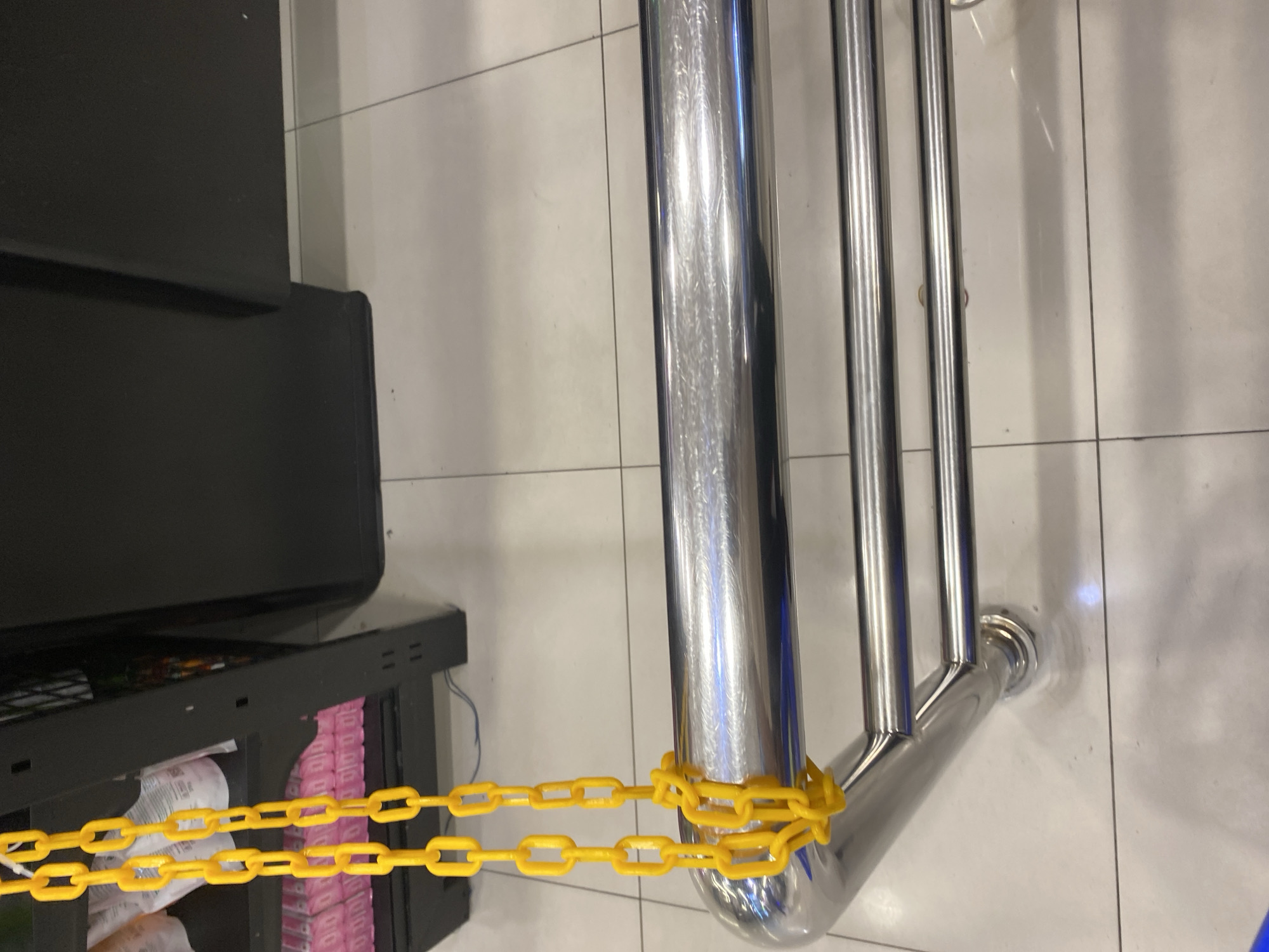 Two yellow plastic chains hang vertically in the foreground, looped around a shiny chrome pipe. The pipe is part of a structure with multiple horizontal bars, suggesting a towel rack or display. In the background, a black shelving unit is visible, with stacked pink and white items on its shelves. The floor is tiled with light-colored squares. The scene appears to be indoors, possibly a retail or public space.