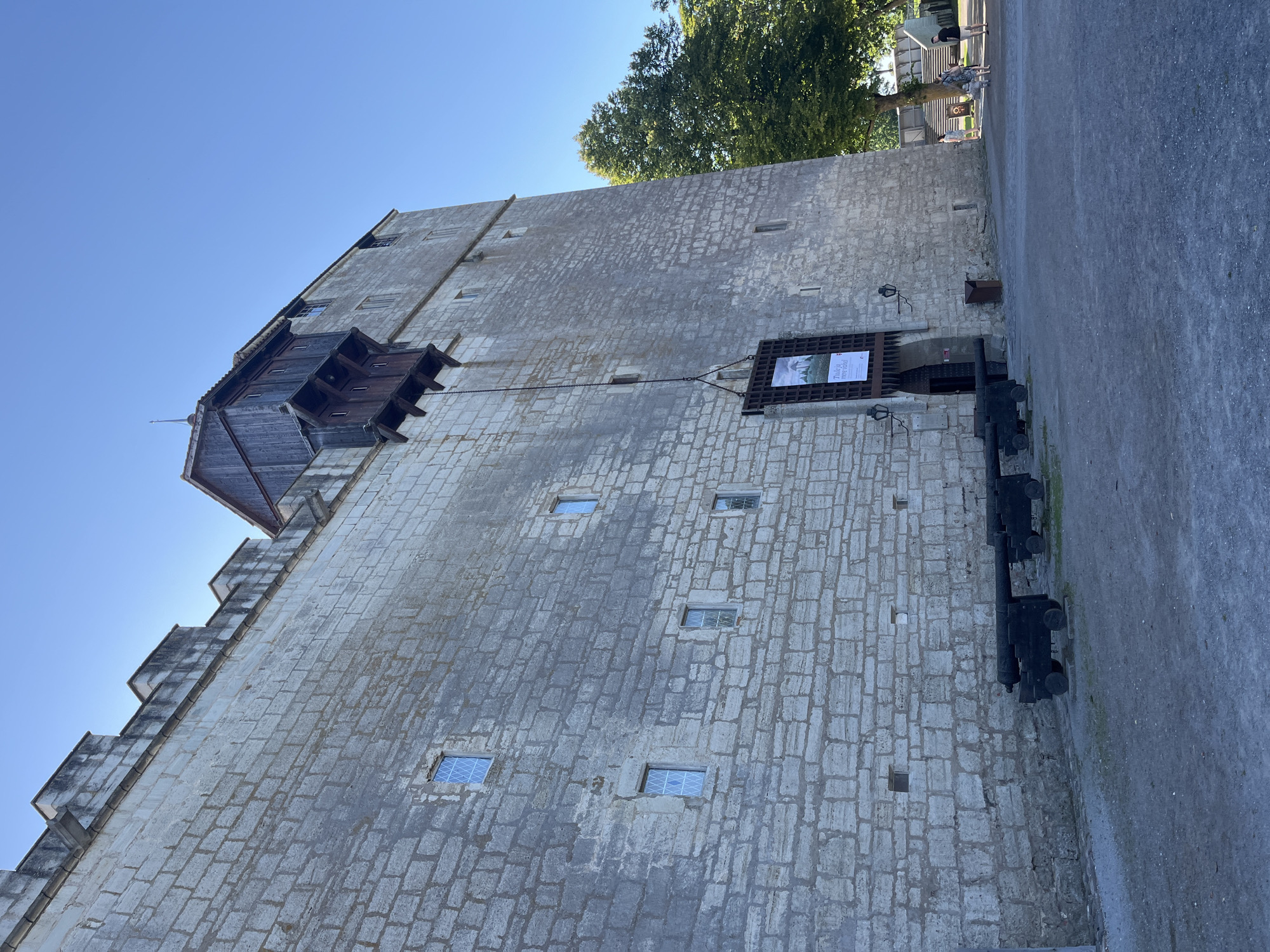The image shows the exterior of Kuressaare Castle in Kuressaare, Estonia, on a clear, sunny day. The castle is a large stone structure with crenellations along the top and a wooden structure projecting from one side. Several small, diamond-paned windows are visible on the stone wall. In front of the castle, two black cannons on wheeled carriages are positioned on the gravel ground. A chain hangs from the wooden structure, connected to a framed poster or banner at the castle entrance. The entrance itself is a large arched opening. To the right of the castle, a large green tree stands, and in the background, a few people are walking near some stairs. The overall scene depicts a historic fortress bathed in sunlight.