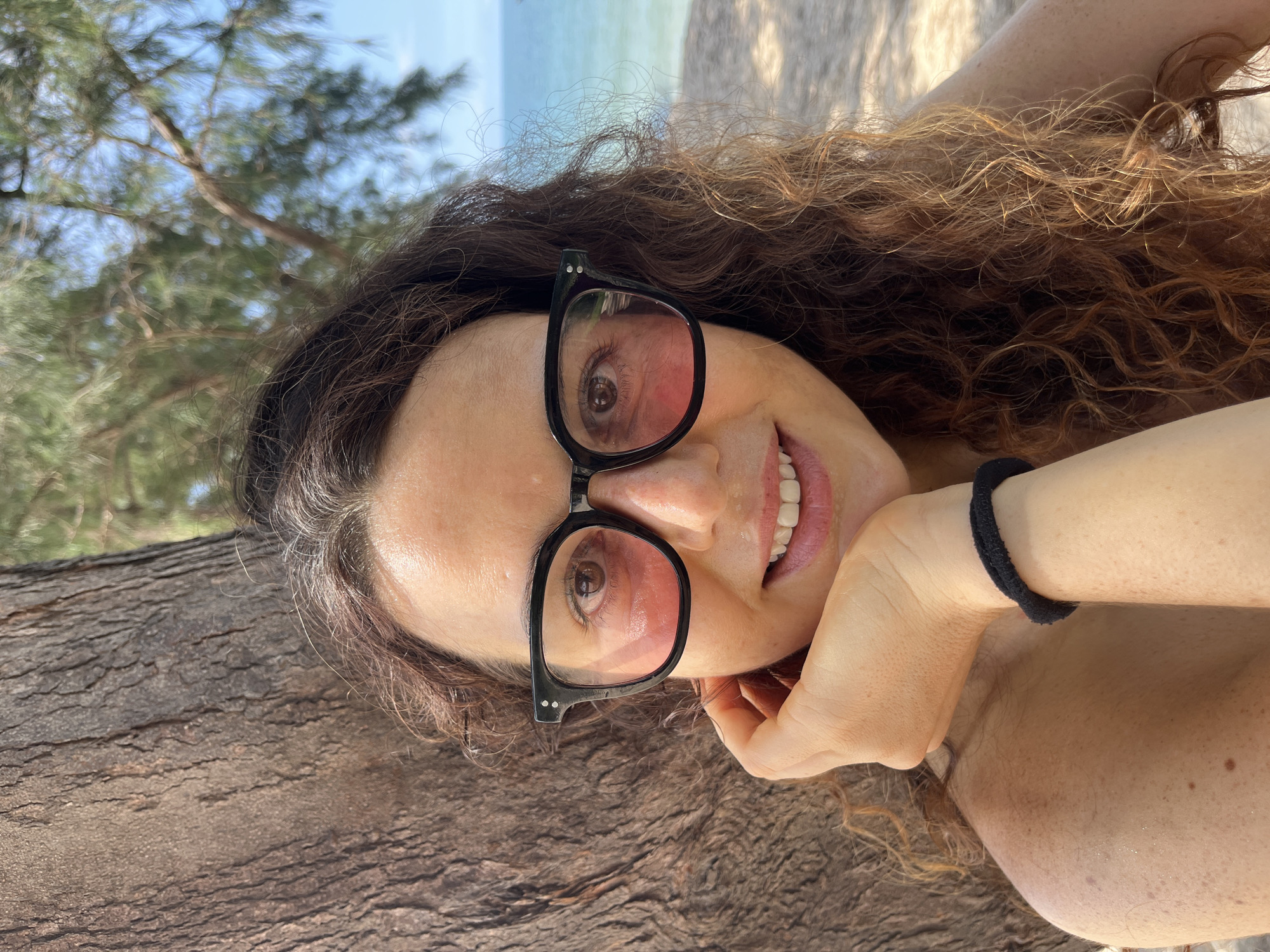 A close-up, selfie-style shot shows a woman resting her chin on her hand, her hair framing her face. She wears black-rimmed glasses with pink lenses and a black hair tie around her wrist. She is outdoors, leaning against a tree trunk, with a beach and the ocean visible in the background. The sunlight illuminates her face and shoulder, highlighting freckles and giving her skin a warm glow. The setting suggests a relaxed, possibly tropical environment.