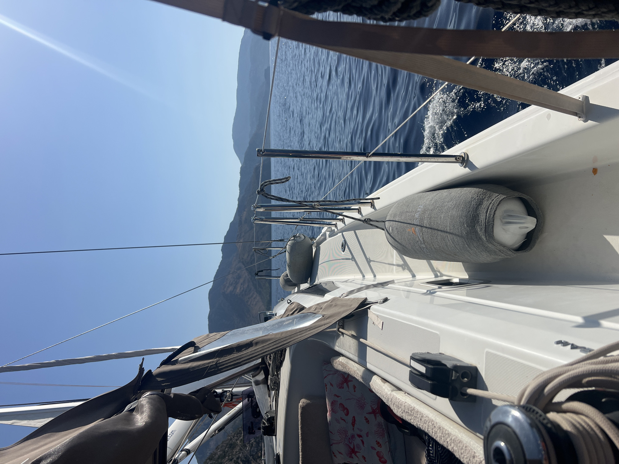 The image depicts a view from the deck of a sailboat moving through calm, blue water. Mountains are visible in the distance, forming a hazy, layered horizon. The sun is shining brightly, creating a lens flare in the upper right quadrant of the image. The deck of the sailboat is white and appears well-maintained, with several nautical features visible. These include a winch, ropes, a cleat, and a grey boat fender. A portion of a sail, partially furled, is visible on the left side of the frame. The water is disturbed by the boat's movement, creating small waves and white foam along the hull. The scene suggests a pleasant day for sailing.