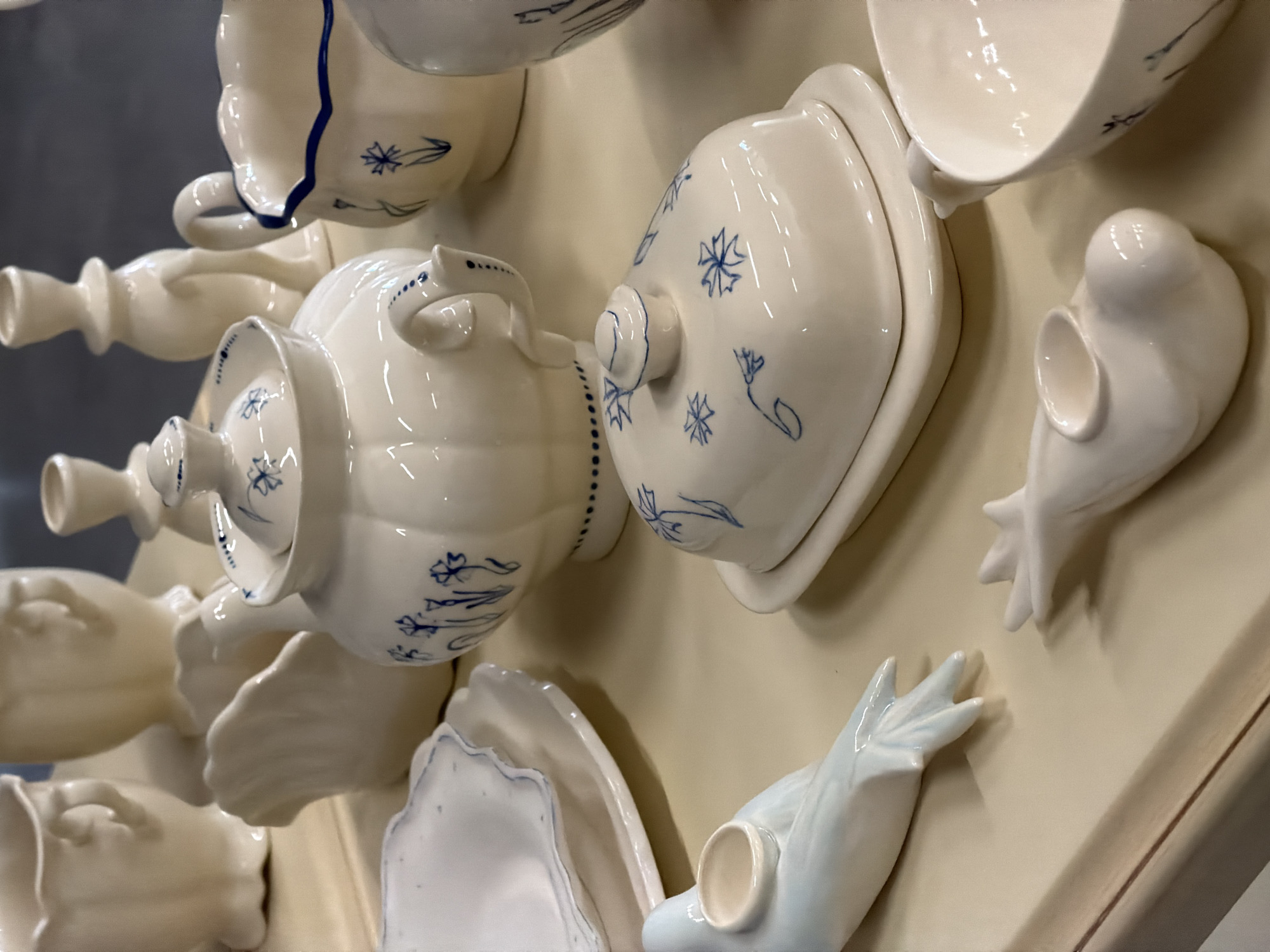 A collection of white ceramic dishware and decorative objects is affixed to a light tan or beige wall or display board. The objects are arranged in a seemingly random, possibly artistic composition. There is a teapot, a covered dish, teacups, decorative candle sticks, scalloped plates and two decorative bird figurines, all executed in a white glaze, some with a delicate blue floral pattern. The background is indistinct, with a hint of a gray wall. The image does not contain information that explicitly identifies the location as Saint Petersburg, Russia, nor does it have details about people, subjects, or interactions. The scene is a static display of ceramics.