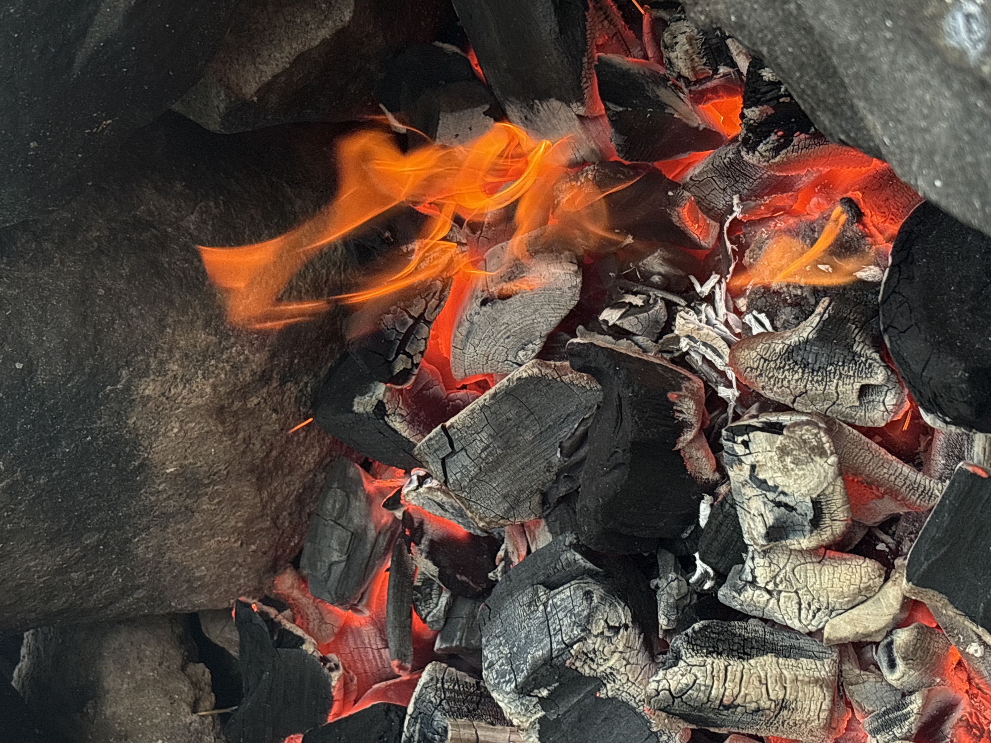 This is a close-up, vertically oriented photograph of a campfire. The scene is dominated by glowing red embers and black charcoal pieces, interspersed with larger, dark grey rocks that frame the fire pit. The flames are a vibrant orange and yellow, licking upwards between the charcoal. The charcoal pieces are varied in size and shape, with some appearing cracked and ashy, indicating they are actively burning. The overall impression is one of intense heat and active combustion. The image does not contain any people or specific identifiable landmarks that would place it in Rautalakhti, Russia, or any other specific city. The scene is entirely focused on the natural elements of the fire and rocks.