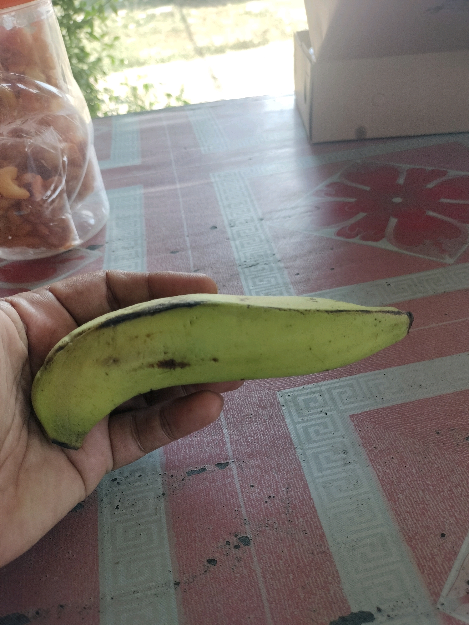 A person's hand holds a single, green plantain on a patterned table. The table has a pink and white striped pattern with a Greek key design. In the background, to the left, is a clear plastic bottle filled with nuts and dried fruit. To the right, a cardboard box sits on the table. The scene appears to be outdoors, with sunlight filtering through foliage in the background. There are no specific indicators of location within Maburai, Indonesia.