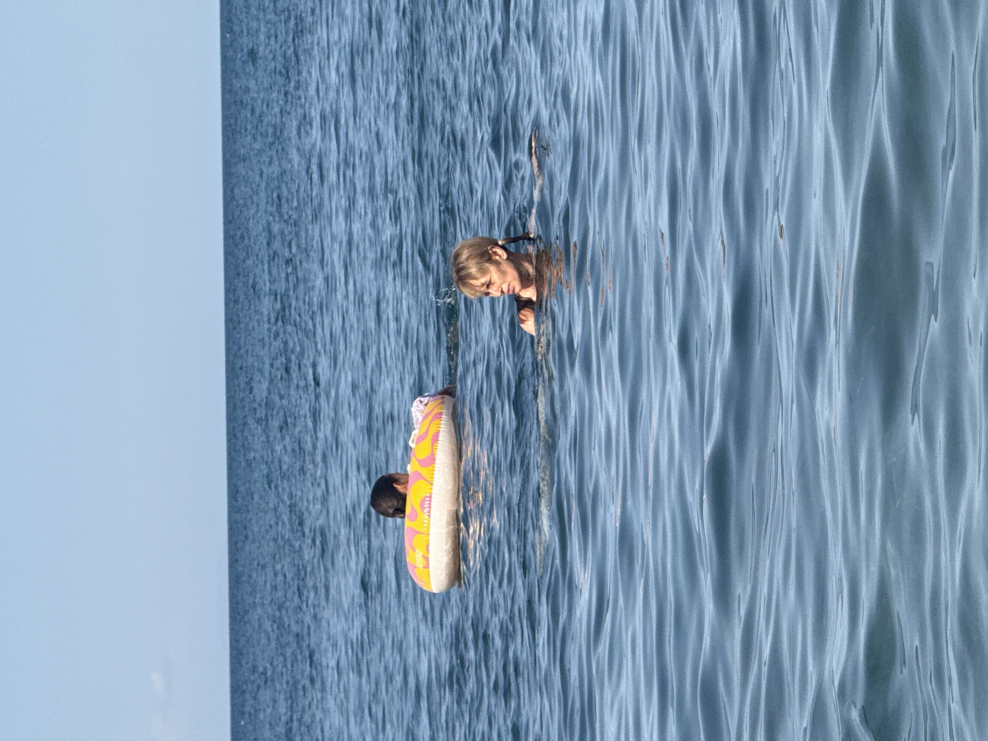 In the city of Partenit, Ukraine, two individuals are observed swimming in the open sea under a clear sky. In the background, a child is partially submerged in the water, utilizing a colorful inflatable ring for buoyancy. The ring displays a pattern of yellow and pink shapes. Closer to the foreground, an adult with short blonde hair is swimming, looking to the left. The water exhibits gentle ripples and a deep blue hue, characteristic of the Black Sea. The horizon line is clearly visible, separating the vast expanse of water from the pale blue sky.