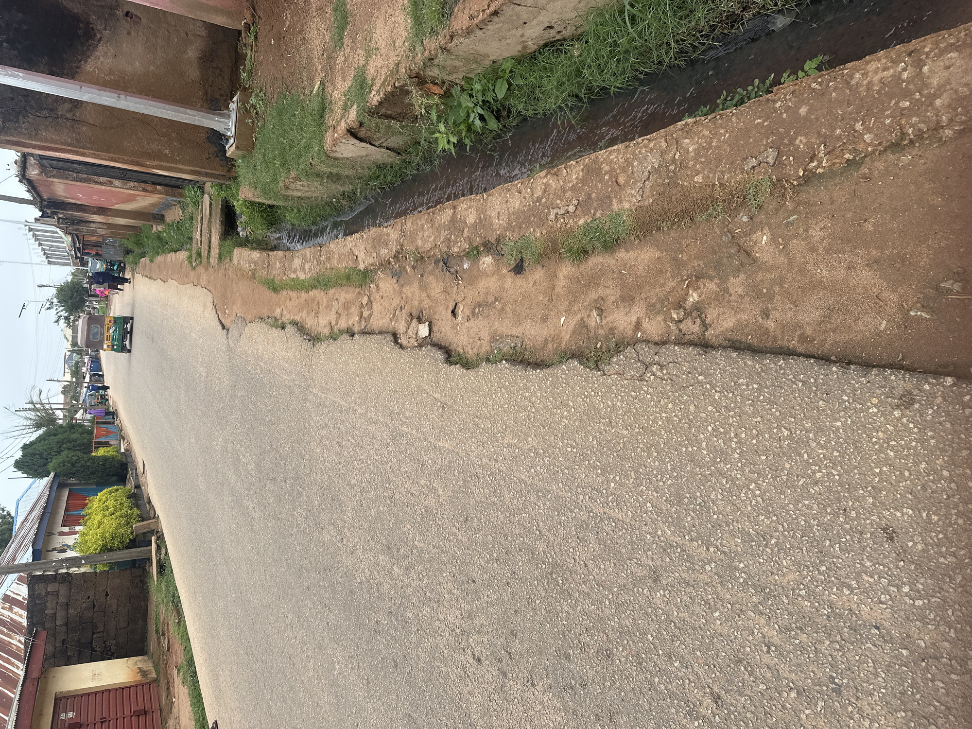 The image shows a street scene in a city. The road is paved with asphalt and appears to be in a state of disrepair, with crumbling edges. To the left of the road, there are buildings with corrugated metal roofs. On the right side of the road, there is a drainage ditch filled with water and overgrown with grass. Several people are walking along the road, and there are a few vehicles, including a three-wheeled auto-rickshaw. The environment appears to be a mixed residential and commercial area. The lighting suggests it is daytime, possibly overcast.