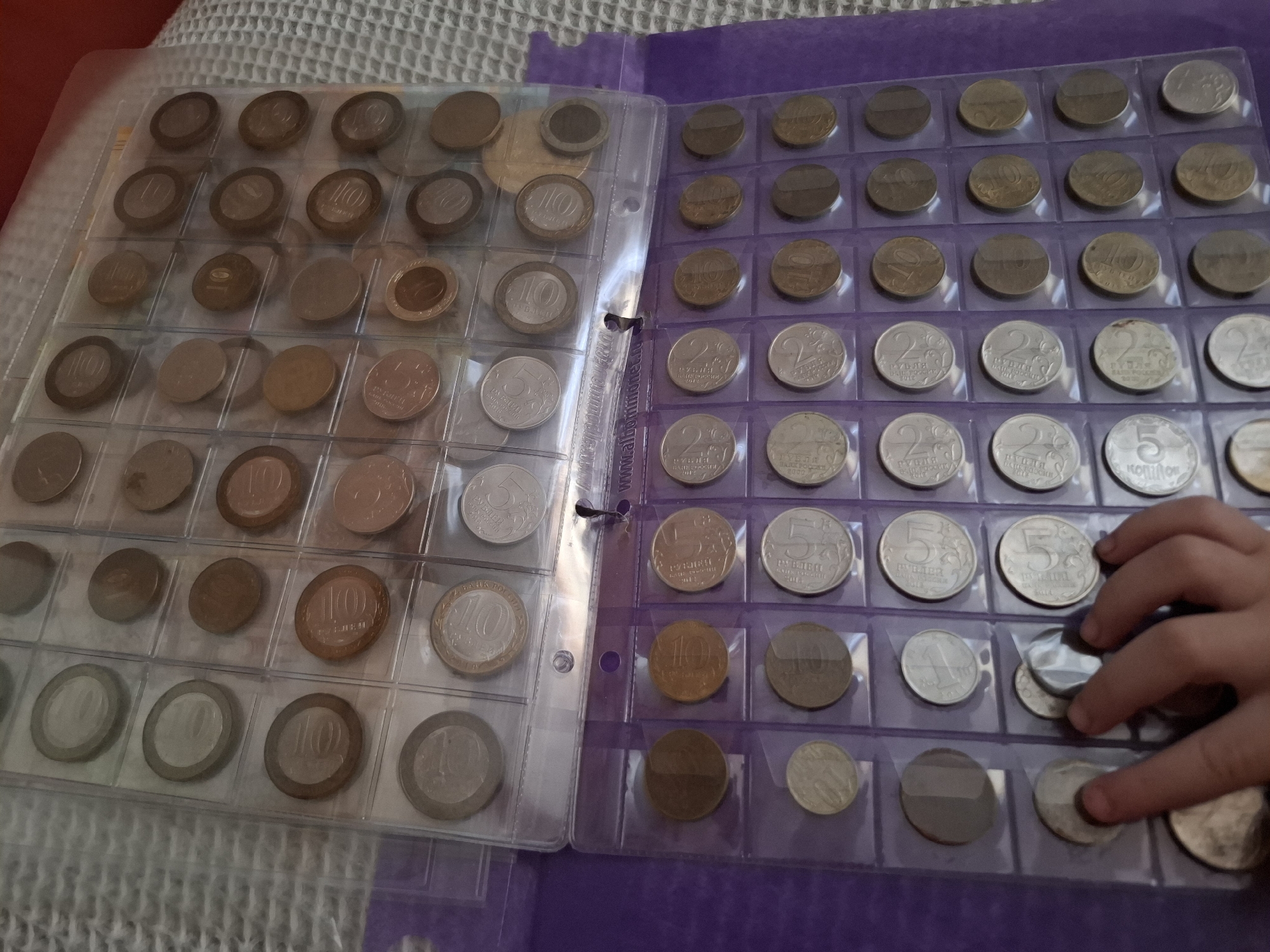 A photograph shows two pages of a coin collecting album. The pages are filled with Russian coins of various denominations, including 10 Rubles, 2 Rubles, and 5 Kopeks. A child's hand is visible on the right page, apparently arranging or examining the coins. The album is resting on a textured surface. The image was taken in Амзя, Russia.