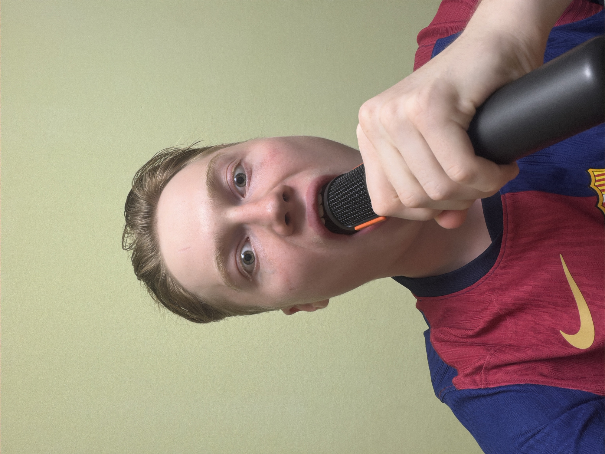 A young person with light brown hair and fair skin holds a black microphone with an orange trim to their open mouth. They are wearing a blue and red striped shirt with a yellow Nike logo. The background is a plain, light green wall. The person's expression is one of surprise or excitement, with wide eyes and an open mouth.