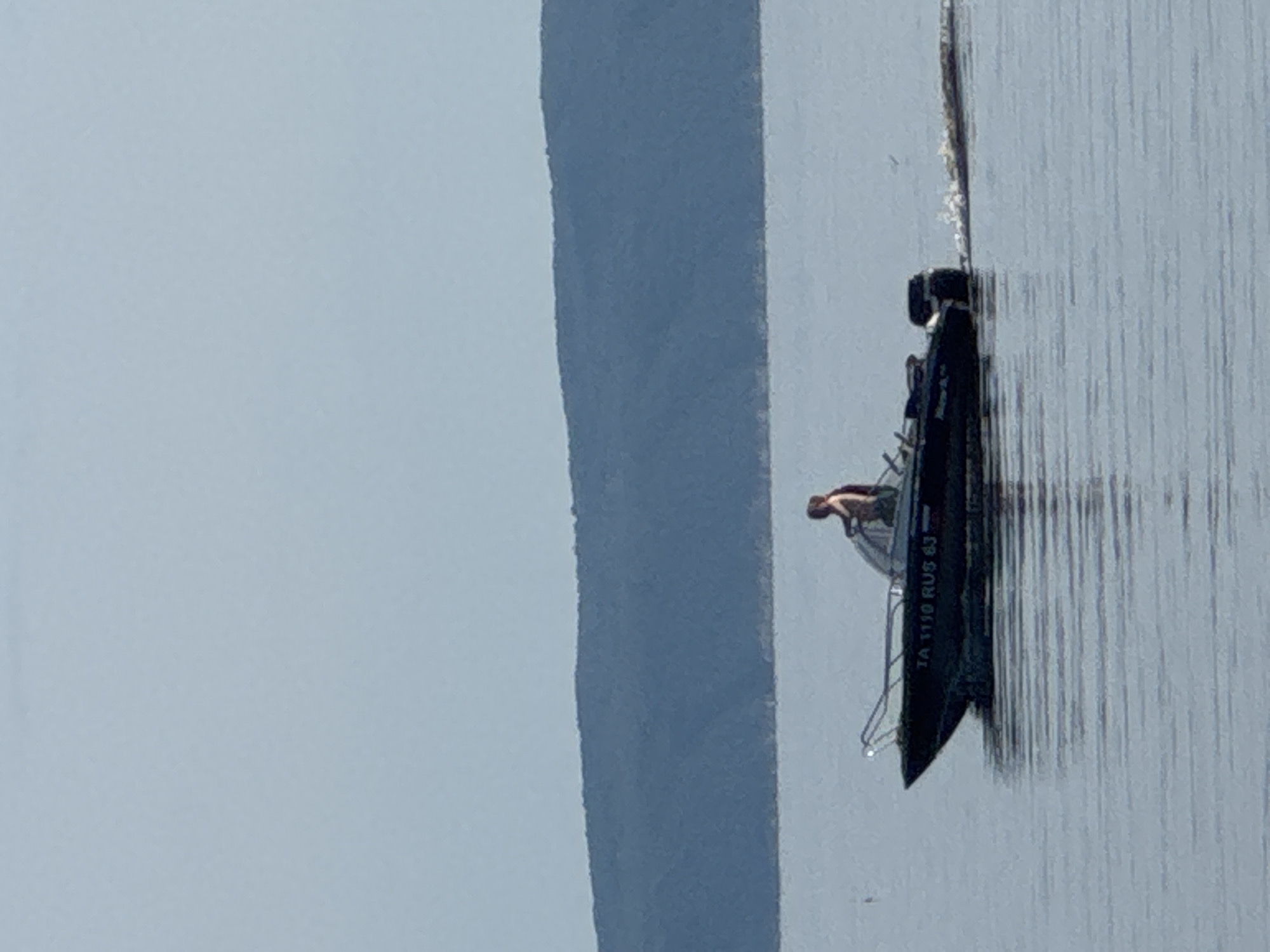 In a calm body of water, a dark-colored motorboat navigates under a pale sky. A lone individual, appearing to be a male, stands at the helm of the boat, his back to the camera. The boat has a visible registration number, "TA 1110 RUS 63", printed on its side. In the background, a wide, dark green, treed hill stretches across the horizon, its base meeting the water. The water's surface is rippled, reflecting the pale sky. The boat is leaving a white wake behind it.
