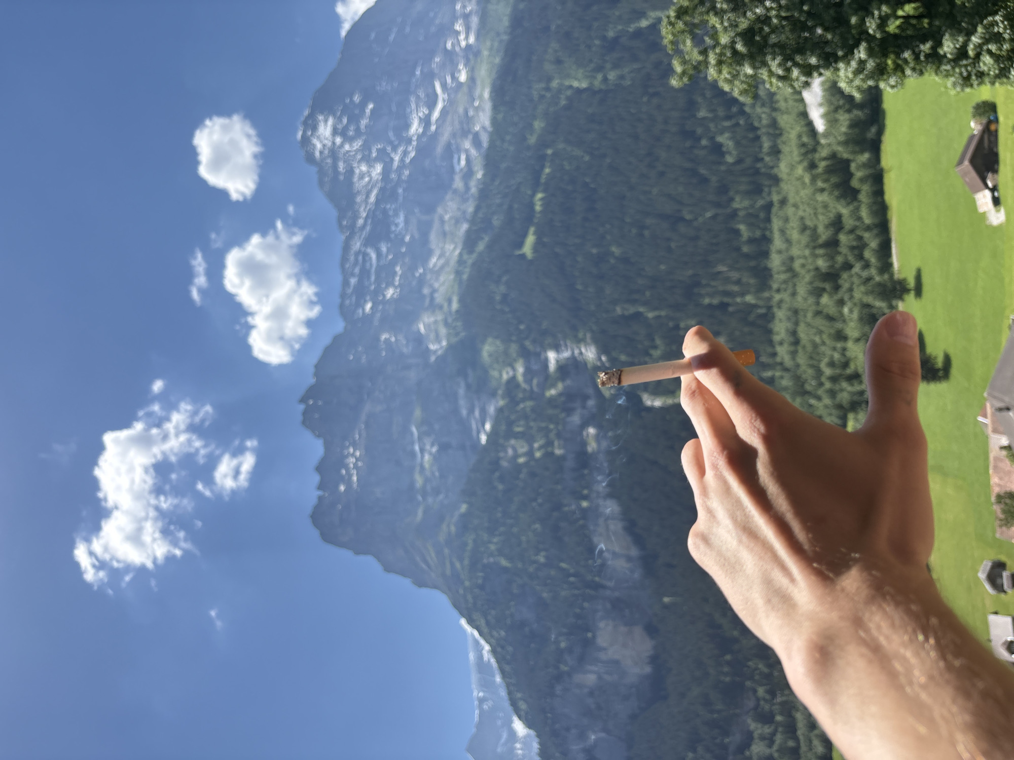 The image depicts a close-up of a person's right hand holding a lit cigarette, with a scenic mountain landscape in the background. The hand is raised, and the cigarette is positioned between the thumb and index finger, with a wisp of smoke rising from the tip.
The background features a vast mountain range, likely in Grindelwald, Switzerland, characterized by steep, rocky slopes partially covered in lush green trees. A clear blue sky with scattered white clouds dominates the upper left portion of the frame. To the right, a bright green meadow is visible, with a few small buildings and scattered trees.
The scene appears to be set during the daytime, with bright sunlight casting shadows and illuminating the landscape, suggesting fair weather. The angle of the image suggests the viewpoint is from a slightly elevated position, possibly a balcony or window. No text is visible in the image. The overall mood is one of relaxation and enjoyment of the natural scenery.