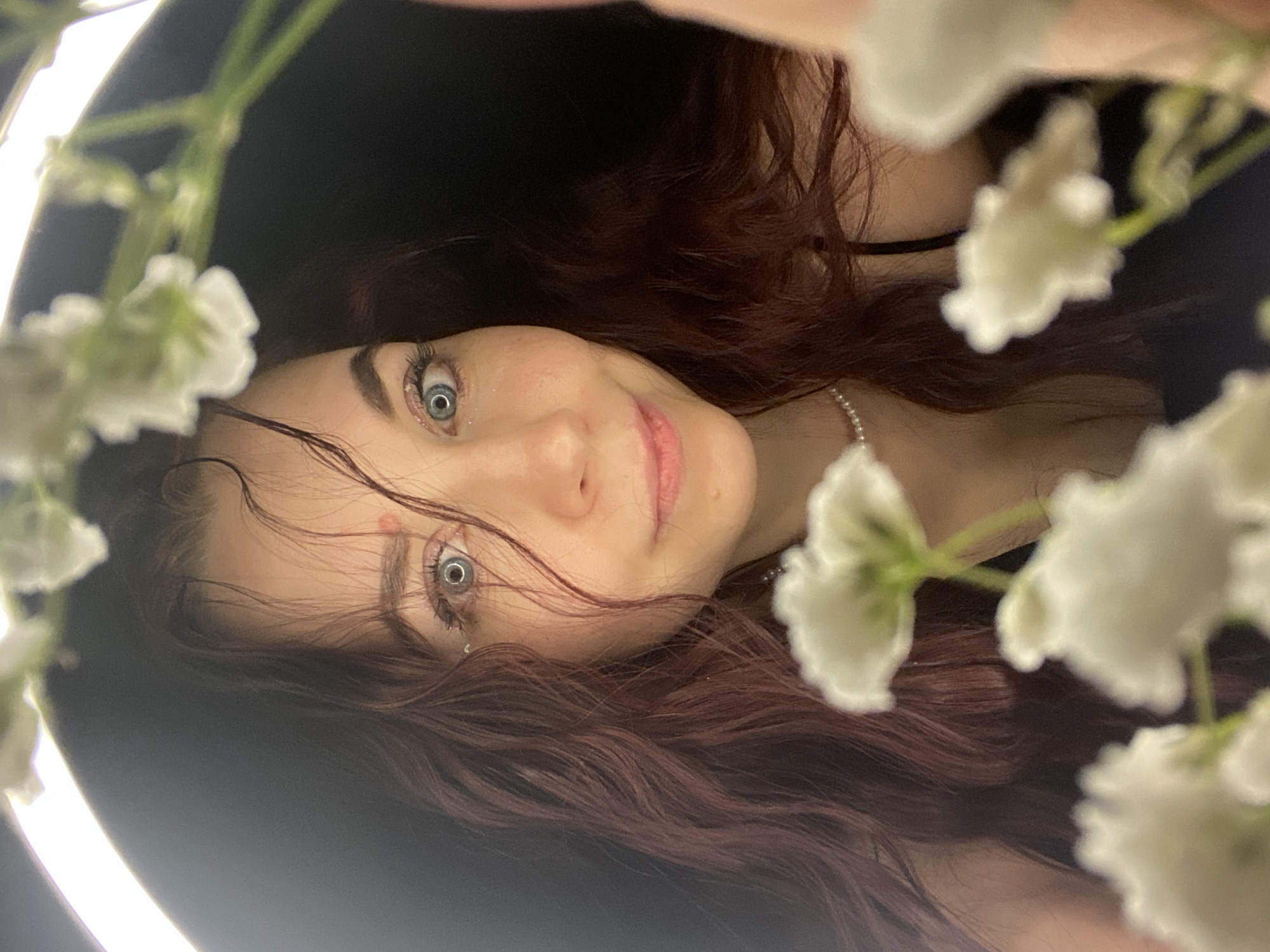 A selfie portrait of a fair-skinned woman with red-brown hair. She has light blue eyes and is wearing a string of pearls around her neck. Her hair is loose and frames her face. She is smiling slightly. Around her face, in the foreground, are sprigs of small white flowers. In the background is a diffused light, likely a ring light. There is no indication of the city of Chaikovsky, Russia.