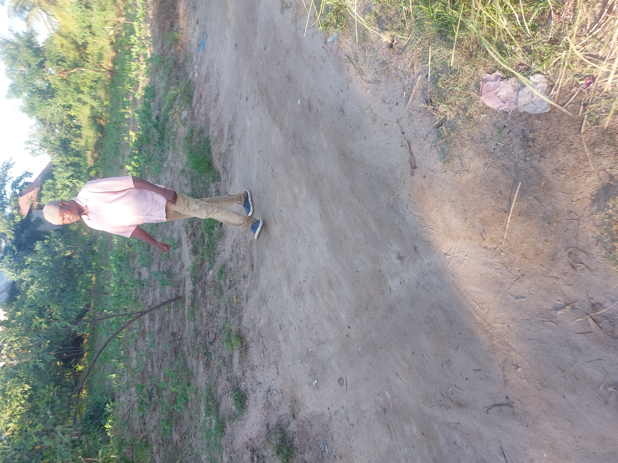 The image depicts an adult Black male standing on a dirt road, likely in a rural or semi-rural area. He is wearing a light pink collared shirt and khaki pants, with blue shoes. He is facing towards the left side of the image, with his left arm extended. The road is dusty and flanked by green vegetation on both sides. The vegetation includes grasses, shrubs, and trees. The sky is bright, suggesting it is daytime. There are some faint shadows on the road, indicating a sunny day. There is no specific information suggesting the location is within the city of Dar es-Salaam, Tanzania, and no specific forensic details are evident.
