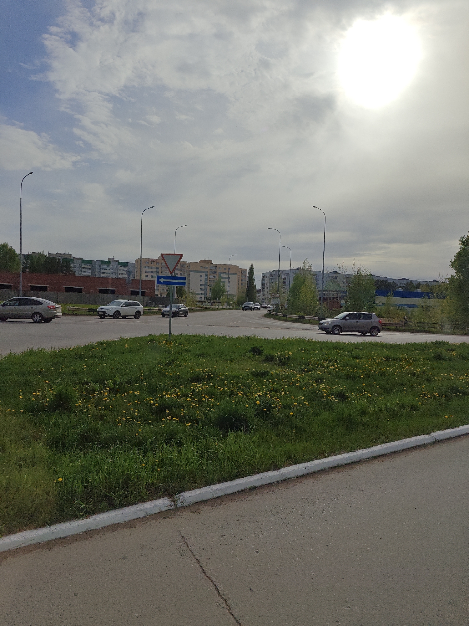 The image captures a scene at a road intersection in the city of Chaikovsky, Russia.
Environment: The day is overcast with the sun obscured by a layer of clouds. The scene features a mix of urban and natural elements, with buildings in the background and green grass populated with yellow dandelions in the foreground.
Objects: The intersection is marked by a yield sign with a blue directional arrow. Several cars are seen driving across the intersection. Tall street lights are installed along the roads. A concrete curb divides the road from the grassy area.
Activities: Cars are actively driving through the intersection.