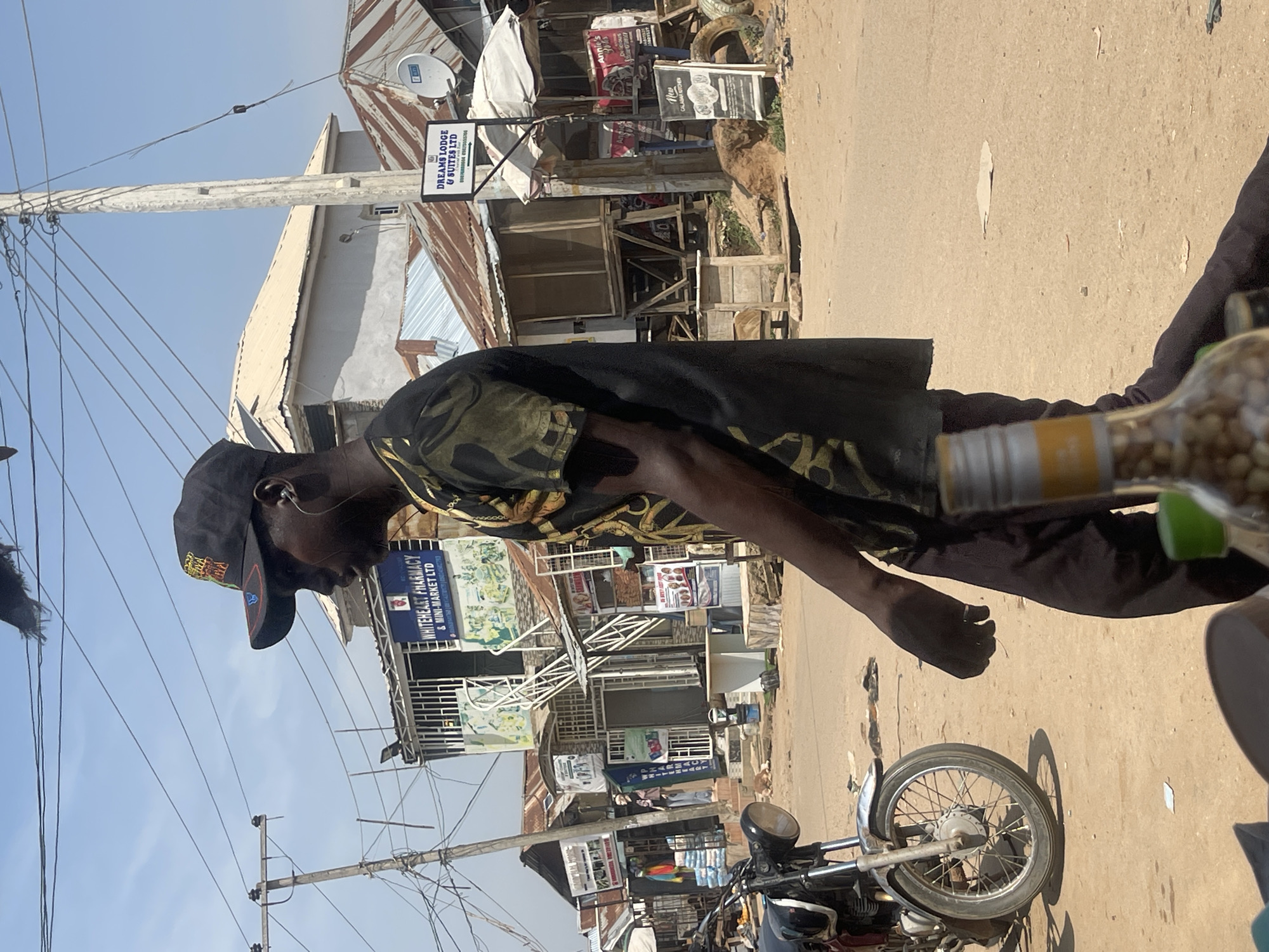 The image depicts a street scene in Abuja, Nigeria, likely in an area with informal markets and businesses. In the foreground, a person wearing a black baseball cap and a dark shirt with a camouflage-like pattern is walking from left to right, partially obscuring a motorcycle parked on the left. The motorcycle appears to be a standard model with a chrome front fender. The background shows a row of shops and stalls with corrugated iron roofs, typical of many urban areas in Nigeria. Signs for "WHITEHEART PHARMACY & MINI-MARKET LTD" and "DREAMS LODGE & SUITES LTD" are visible, indicating commercial activity. Utility poles with numerous wires crisscross the sky, suggesting a developed but perhaps aging infrastructure. The ground is paved with dirt and loose stones, with some litter visible. A partially consumed bottle with a yellow label is in the lower right corner of the frame, out of focus. The overall impression is of a bustling, everyday street in a Nigerian city.