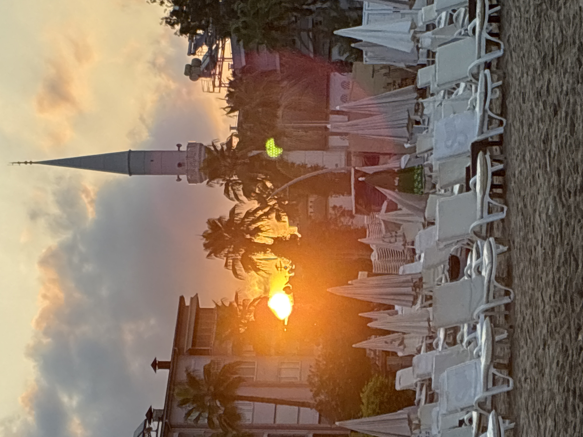 The image shows a beach scene at sunset in Alanya, Turkey, with the sun setting behind palm trees and buildings. In the foreground, there are rows of white lounge chairs and closed beach umbrellas on the sand. Behind them, there is a building with several floors and balconies, and a tall minaret of a mosque. The sky is filled with dramatic clouds, illuminated by the orange glow of the setting sun. Palm trees are scattered throughout the scene. The overall atmosphere is peaceful and serene, with a hint of warmth from the sunset.