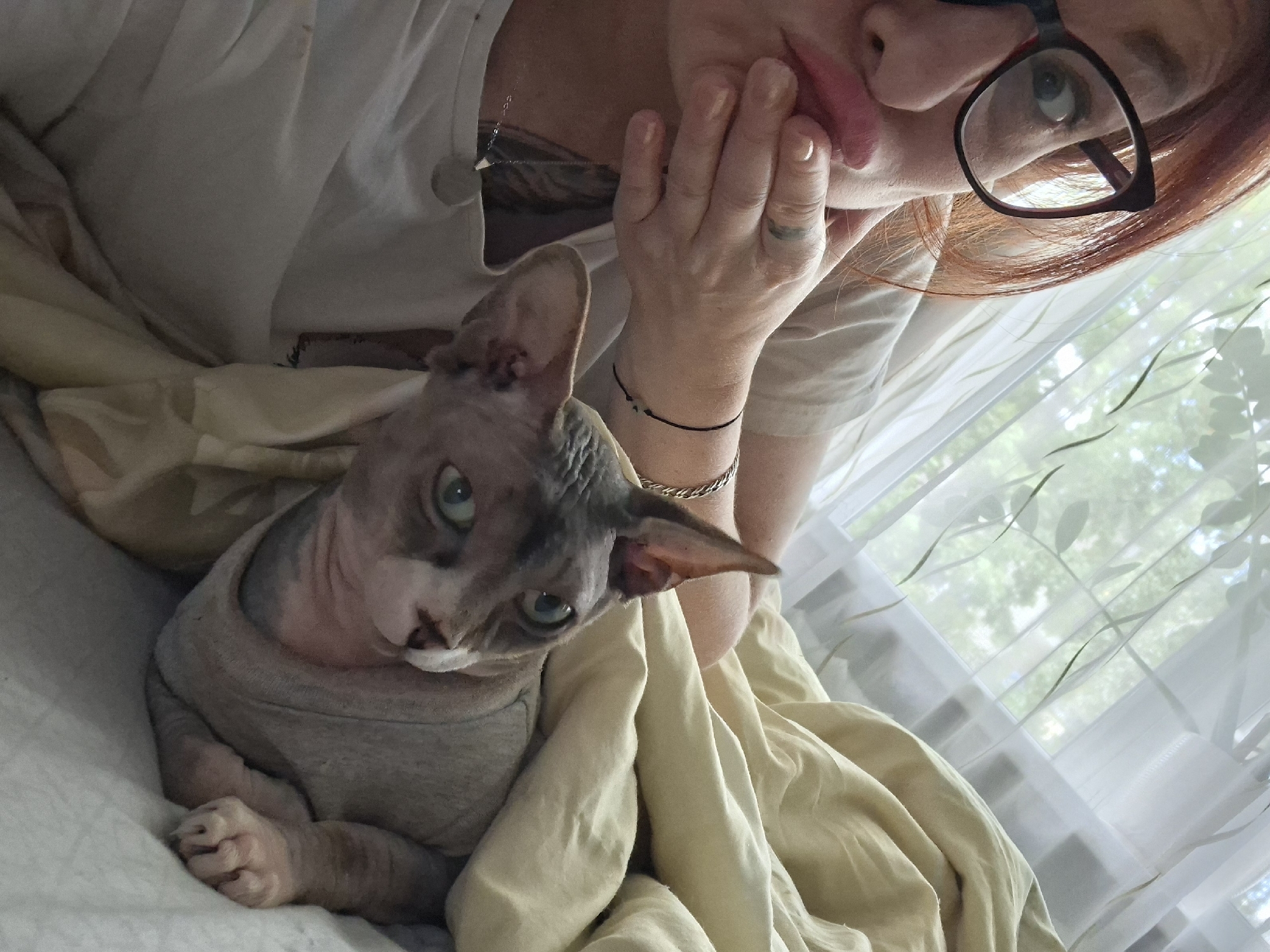 A woman with red hair, wearing black-rimmed glasses, is lying in bed with a Sphynx cat. She is looking at the camera with her hand covering her mouth. The cat is partially covered by a beige blanket and is wearing a grey shirt. The background shows a window with sheer curtains and greenery visible outside. The scene appears to be indoors, likely a bedroom. The location is not specified to be Myadzyel, Belarus.
