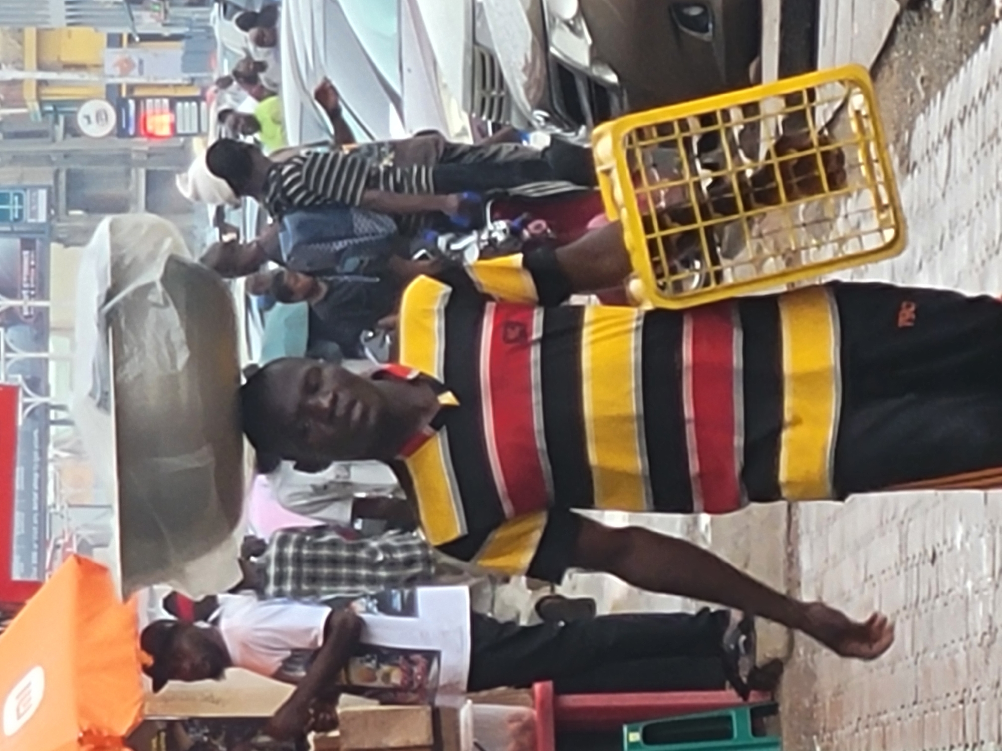 A man is walking in the city of Jos, Nigeria. He is wearing a horizontally-striped shirt in black, yellow, and red, with black shorts. He is carrying a yellow plastic crate on his shoulder. Other people are walking on the sidewalk and near vehicles, some are visible in the background.