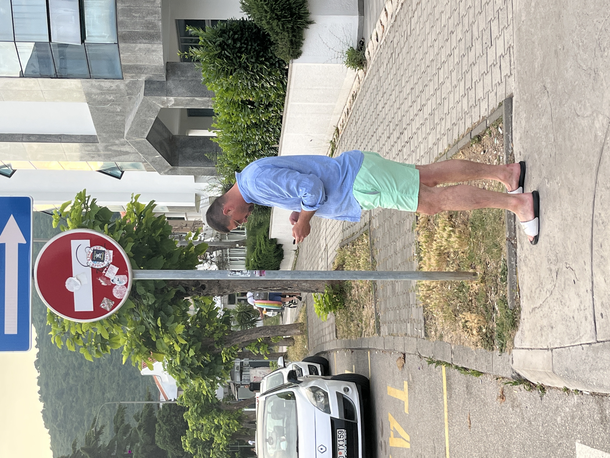 A man stands on a sidewalk, looking at his phone. He is wearing a blue button-down shirt and light green shorts, with white slides on his feet. Behind him, a white car is parked on the street. A red and white "no entry" sign is mounted on a pole to his left, adorned with several stickers. In the background, a modern building with glass windows and a lush green hill are visible. The scene appears to be in an urban environment, possibly a resort town, with a mix of residential and commercial structures.