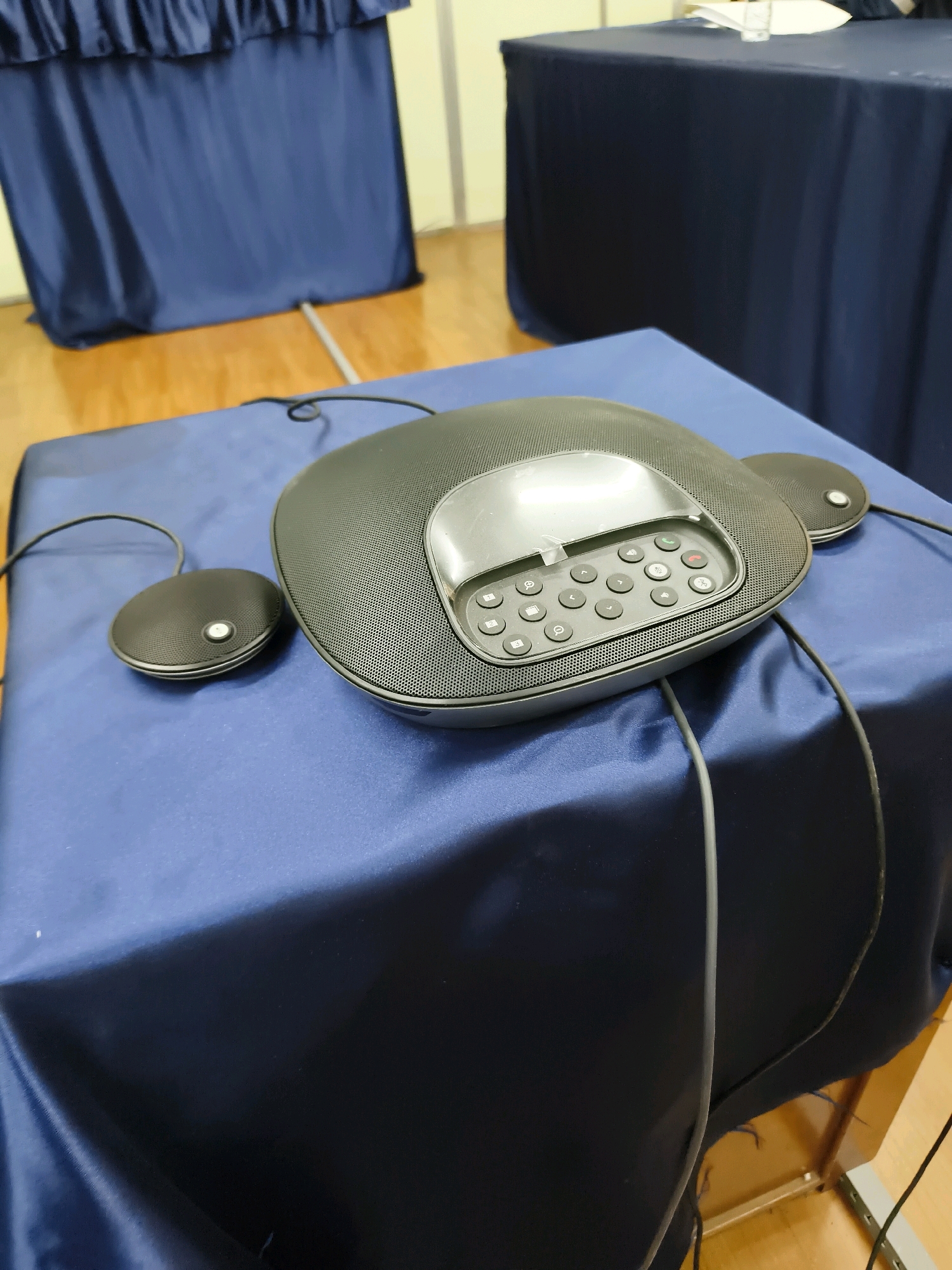A black conference call speakerphone unit with two attached microphones sits on a table covered with a dark blue tablecloth. The speakerphone unit has a keypad with several buttons, including green and red call buttons, and a small screen. The two microphones are circular and connected to the main unit by wires. The table is positioned in front of a dark blue curtain. In the background, another table covered with a dark blue tablecloth is visible, and a light-colored wall and wooden floor can be seen. The scene is indoors, likely in a meeting room or conference space. The image was taken at an angle looking down at the speakerphone.