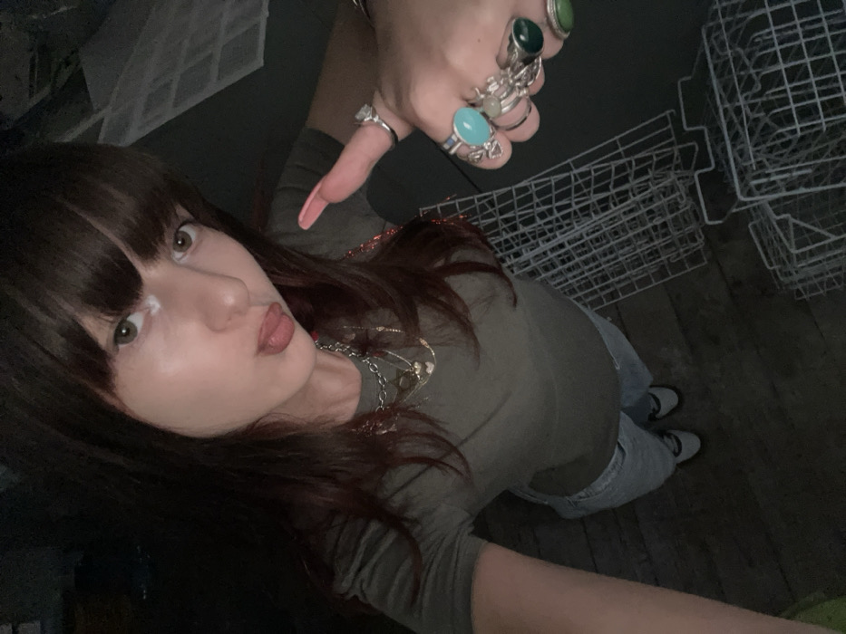 A high-angle photograph depicts a female subject in a dimly lit indoor environment within the city of Raanana, Israel. The subject has dark brown hair with bangs, light skin, and light brown eyes, with a visible white smudge on the left cheekbone beneath the eye. She is dressed in an olive green short-sleeved t-shirt, light-colored ripped jeans, and dark shoes with white accents. Multiple silver-colored layered necklaces with pendants are visible around her neck. The subject's lips are pursed with the tongue slightly extended, and she looks directly at the camera. Her right hand, visible on the left side of the frame, is raised in a "thumbs up" gesture. The thumb bears a long, pointed, light pink artificial nail. Several silver-colored rings are worn on the fingers of the right hand, including a large clear stone on the index finger, a prominent oval turquoise-colored stone on the middle finger, and two rings with green stones on the ring and pinky fingers. The background, to the subject's right, features several stacked white wire grid panels or shelving units. The floor beneath the subject appears dark and textured. The scene is illuminated by dim, directional light.