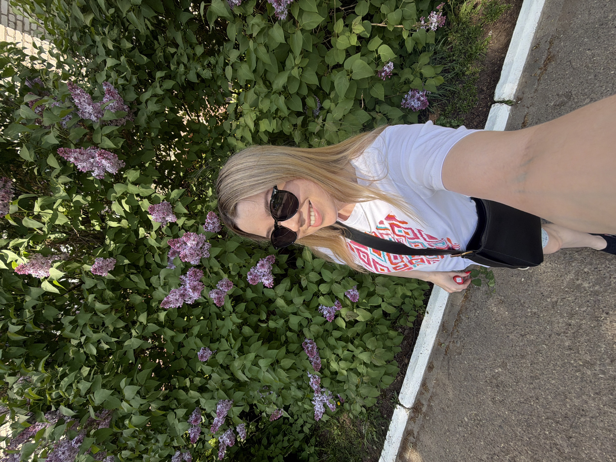 A woman with long blonde hair is posing for a selfie in the city of Chaikovsky, Russia. She is wearing sunglasses and a white t-shirt with a colorful design. She has a black purse slung over her shoulder. The woman is standing next to a lilac bush with purple flowers. The background includes a sidewalk and a white curb.